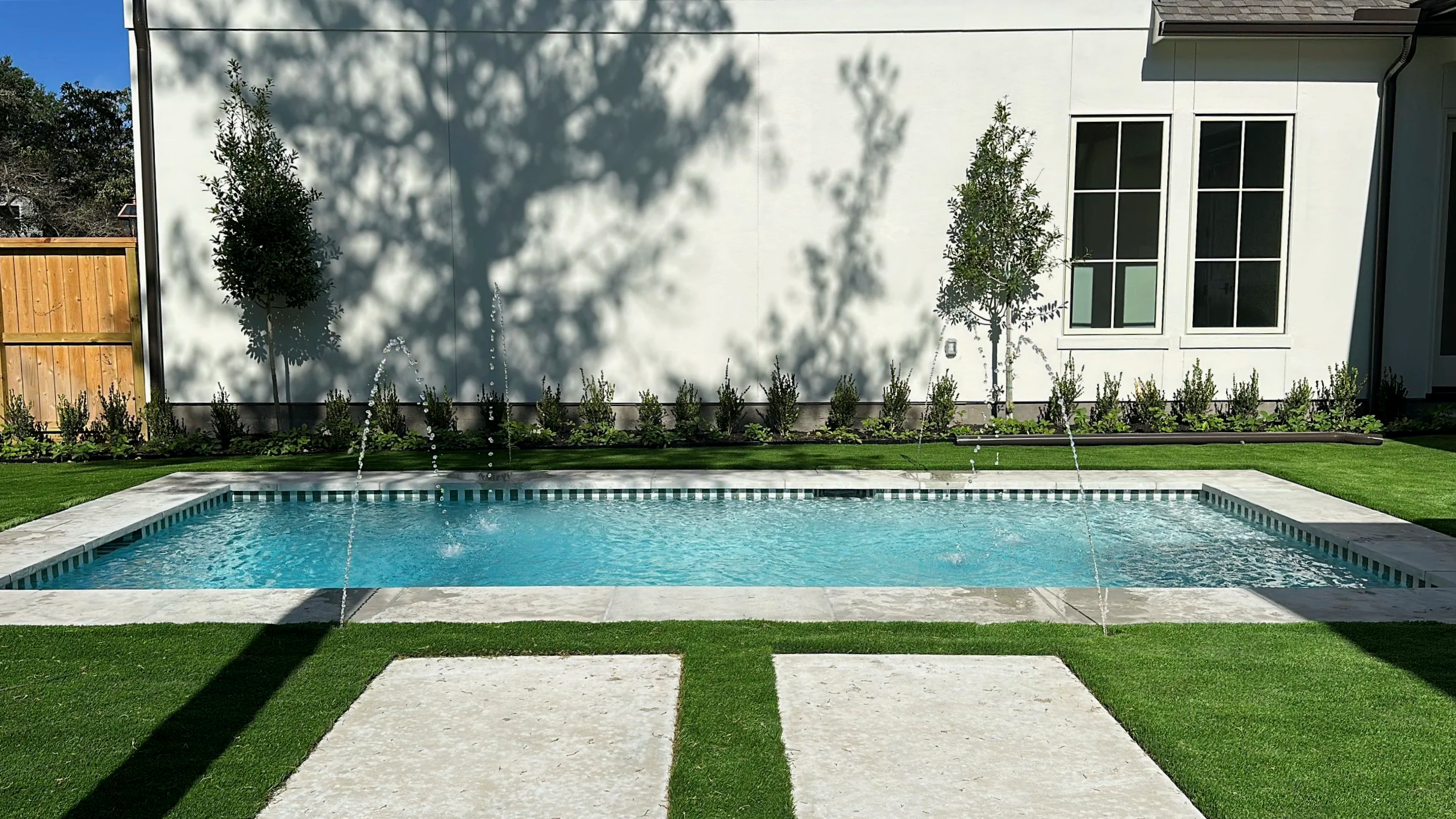 Backyard with a rectangular swimming pool with water fountains, surrounded by green grass, a patio, and a white house with large windows. There are trees casting shadows on the wall.