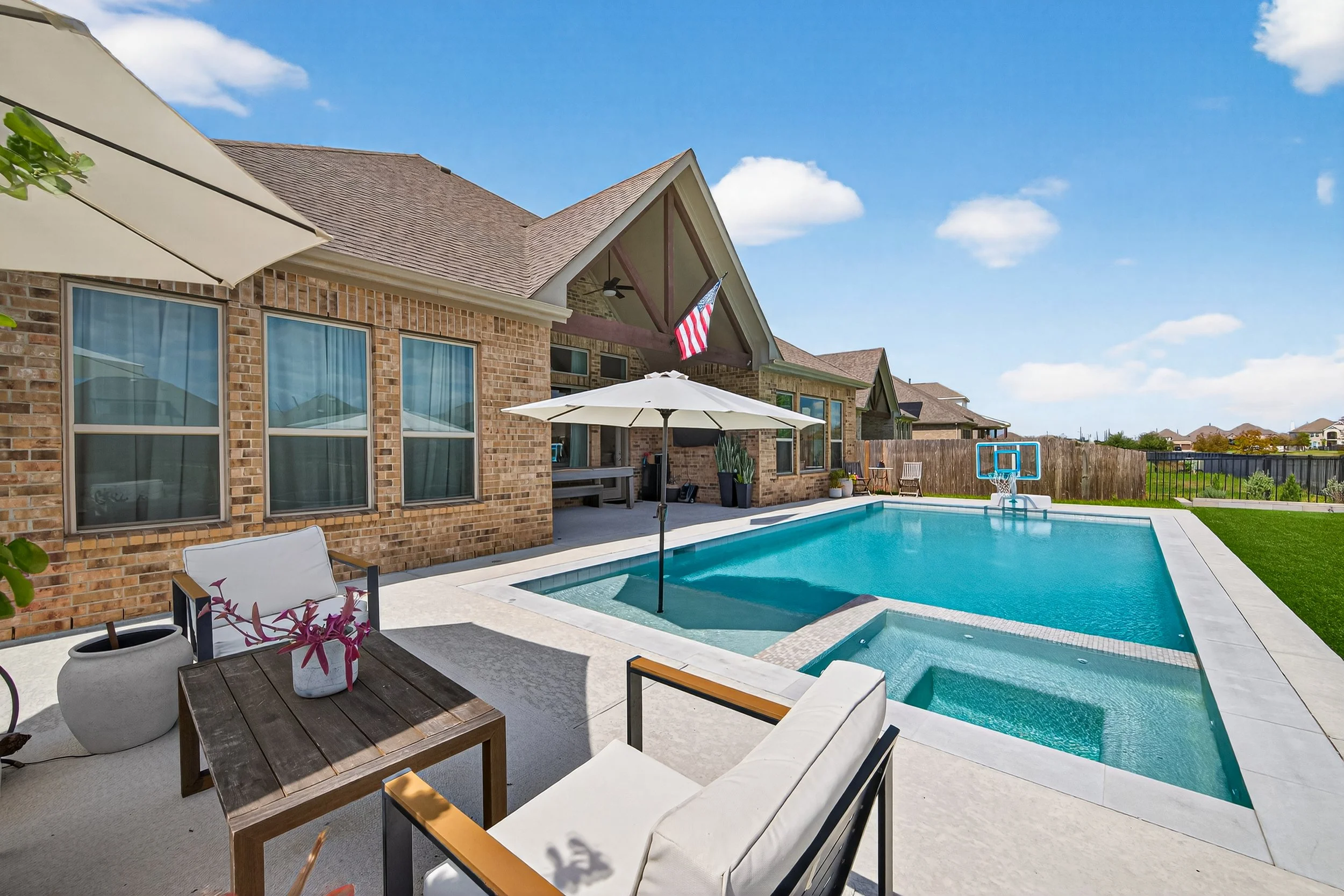 Backyard with a swimming pool, patio furniture, an umbrella, and a basketball hoop, attached to a brick house with large windows under a blue sky.