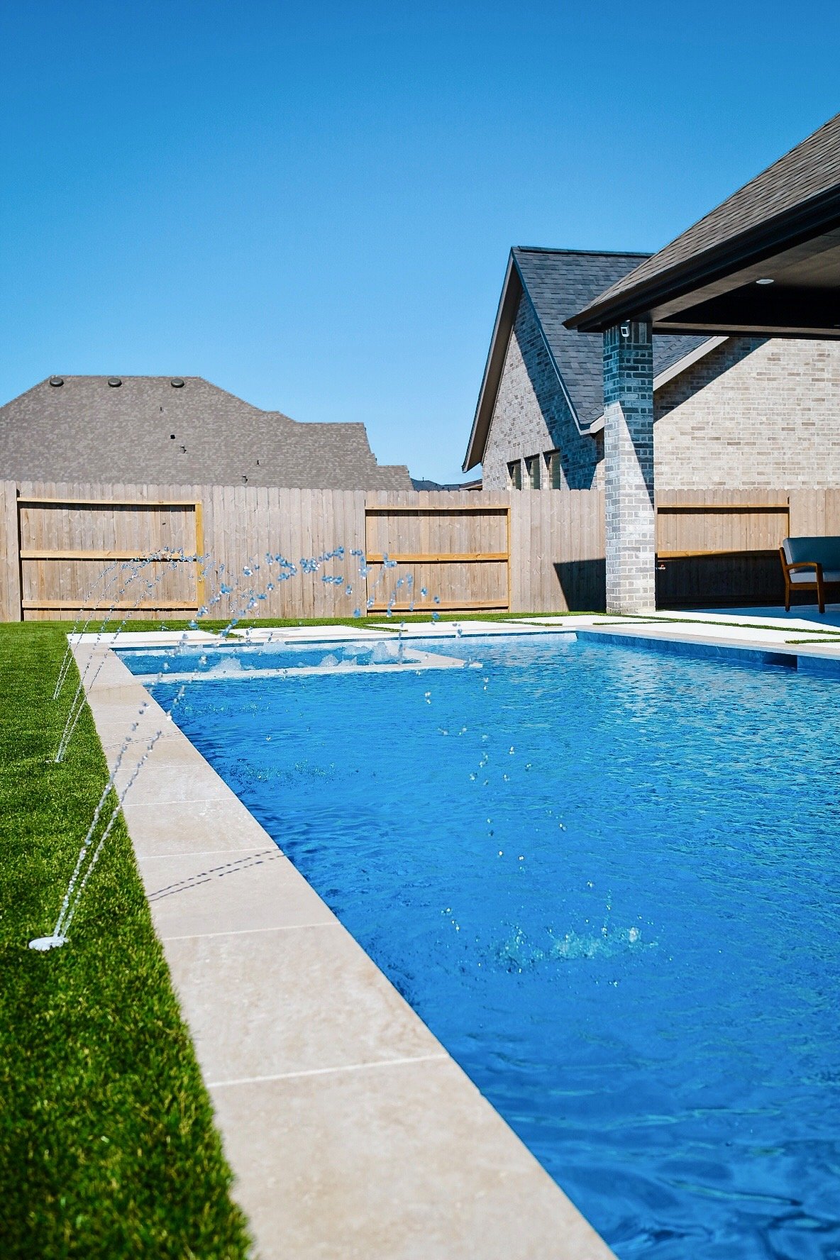 A backyard with a blue swimming pool, small fountain jets, a grass lawn, a wooden fence, and nearby houses under a clear blue sky.