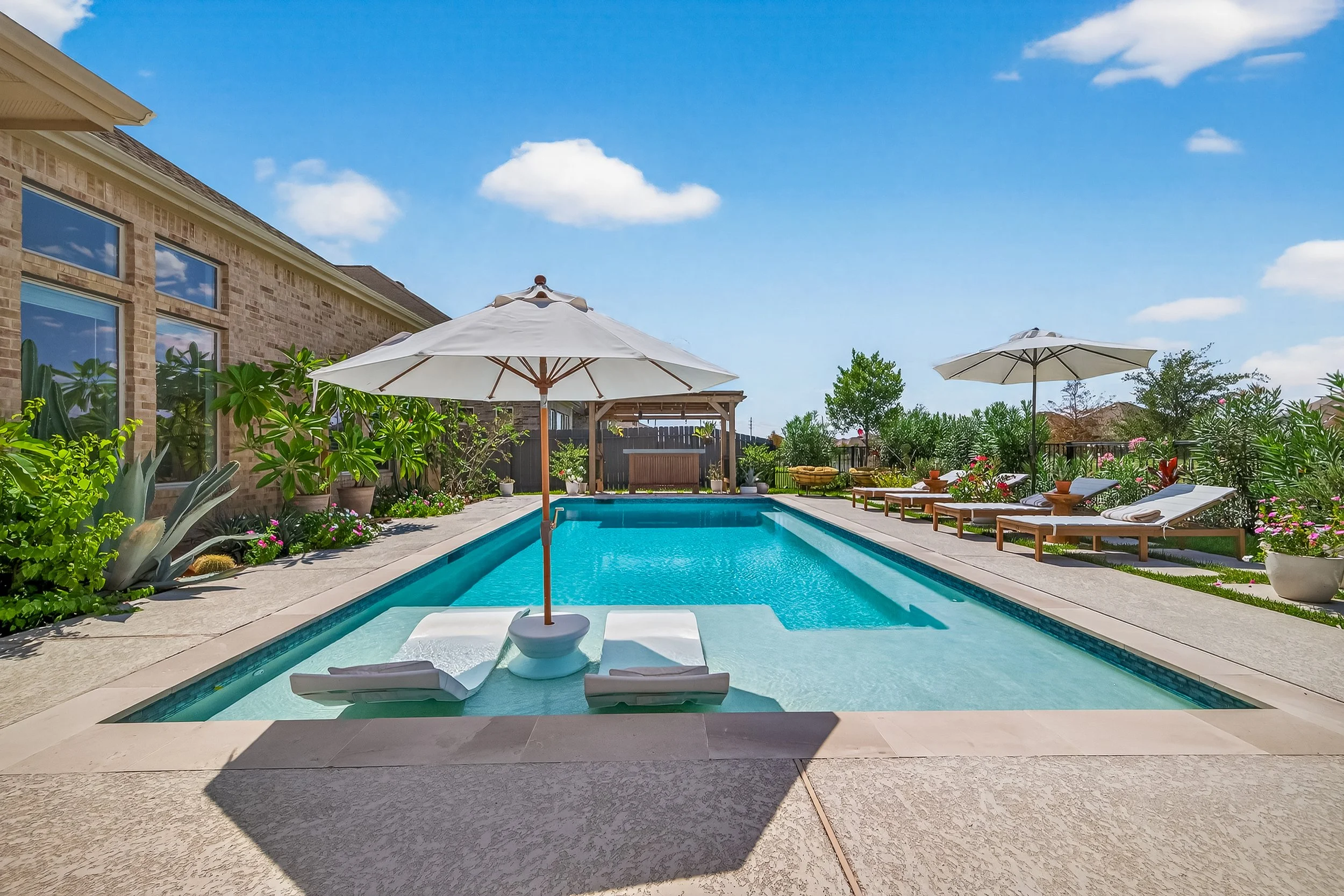 A backyard swimming pool area with two white lounge chairs under a large white umbrella, surrounded by lush green plants, colorful flowers, and additional lounge chairs with umbrellas on a bright sunny day.
