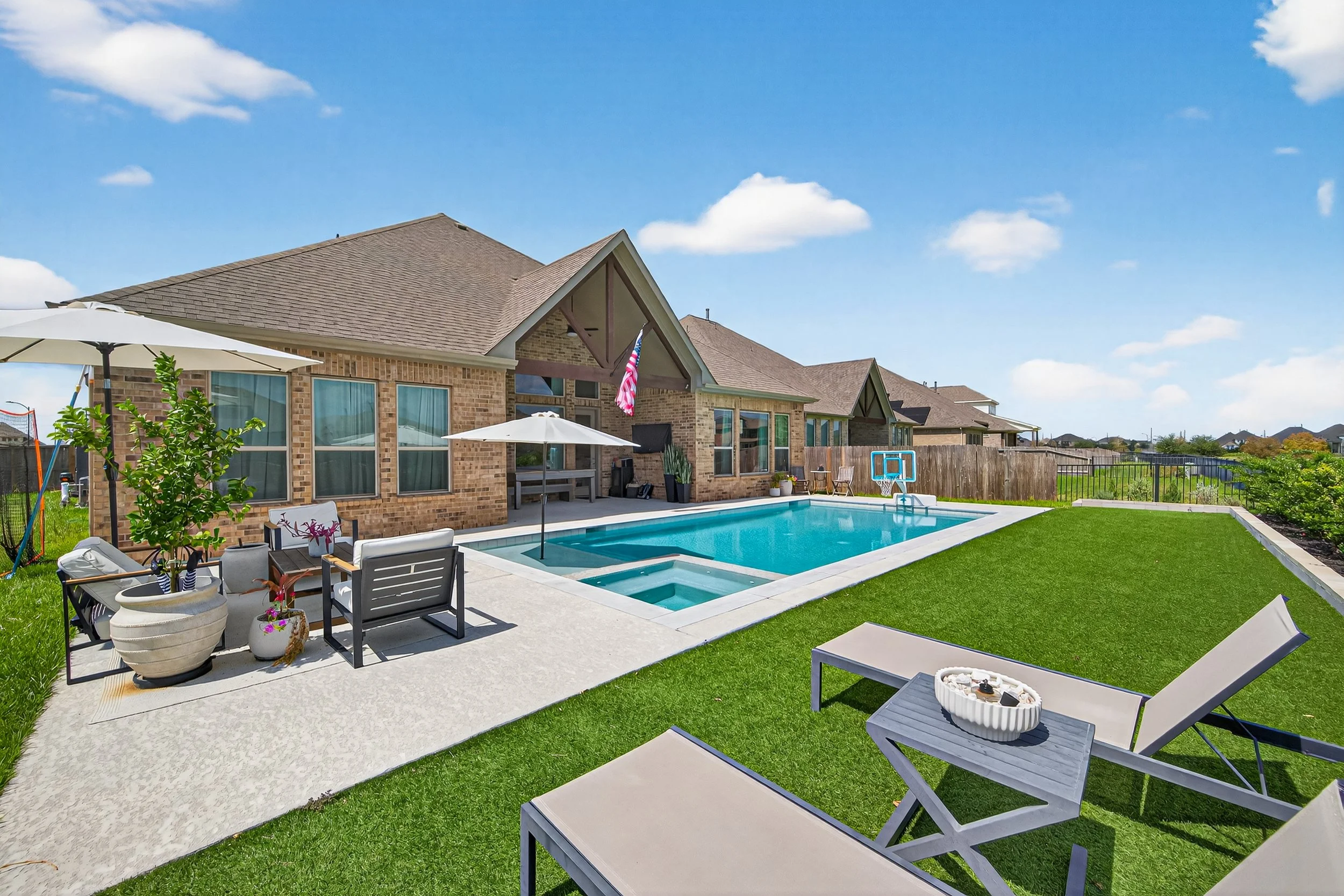 Backyard with swimming pool, lounge chairs, umbrellas, and patio furniture, adjacent to brick house with large windows and a American flag.