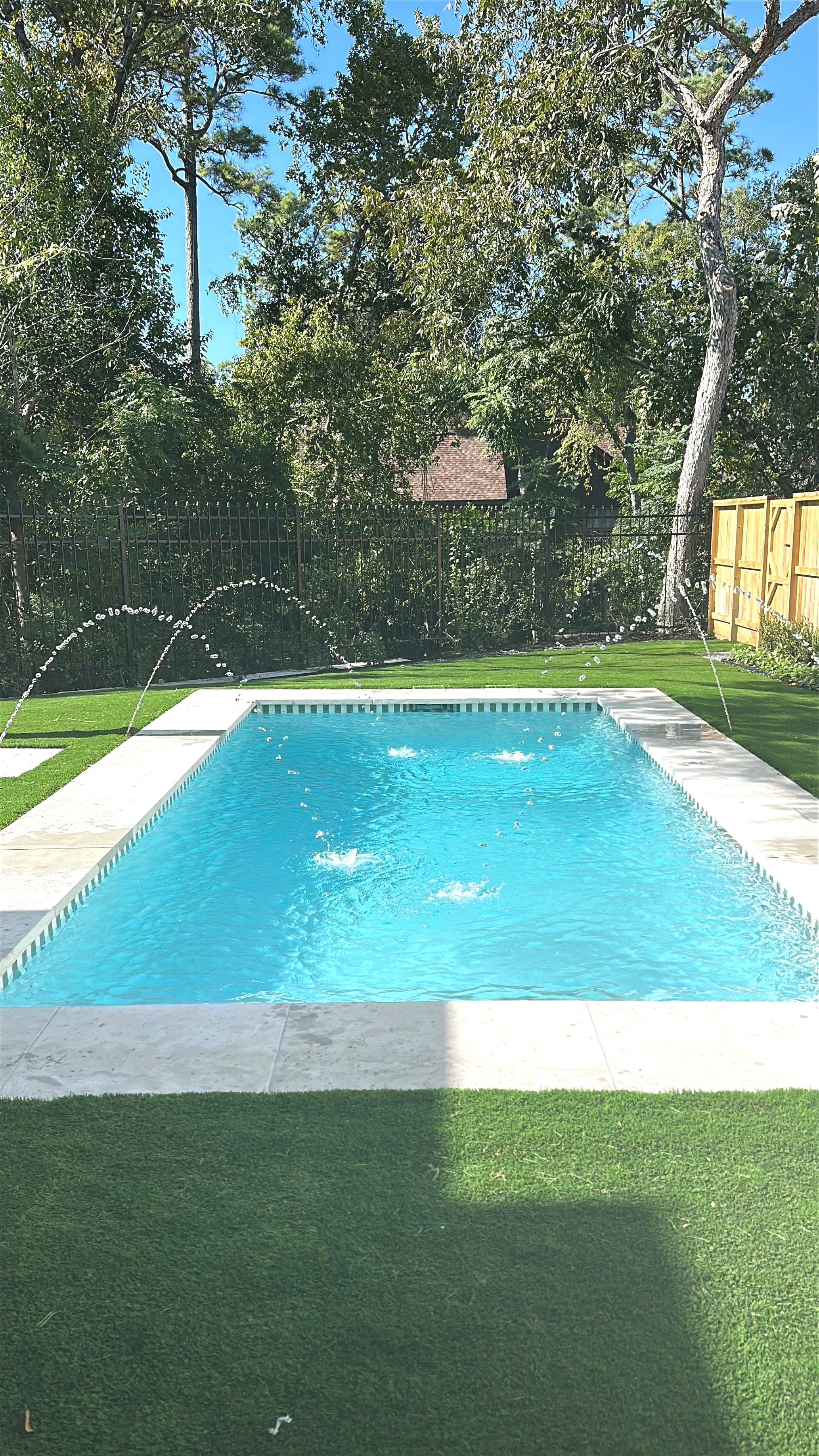A backyard swimming pool with water fountains, surrounded by artificial grass and trees.