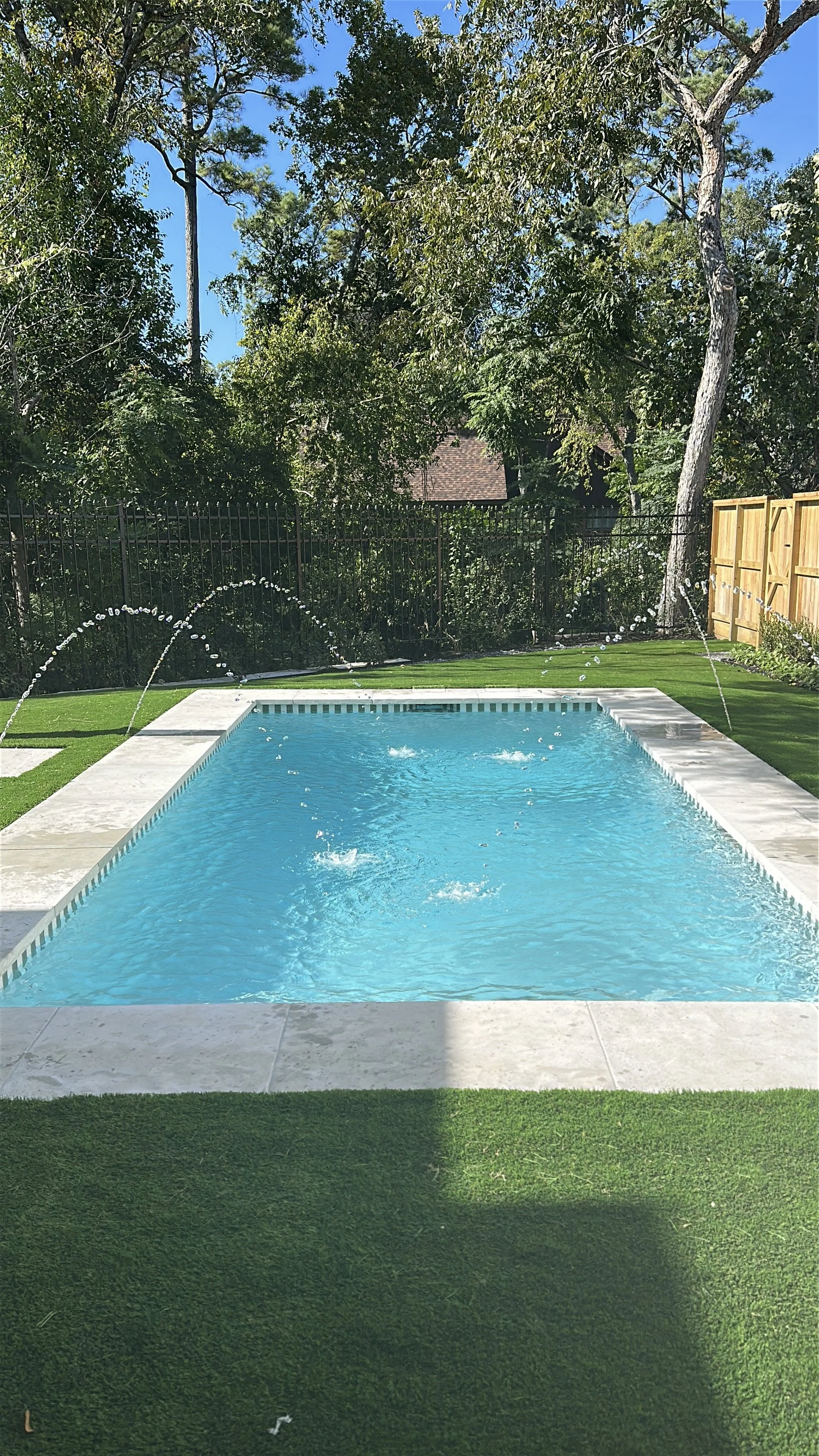 Backyard swimming pool with sprinkler jets shooting water, surrounded by grass and trees, under a clear blue sky.