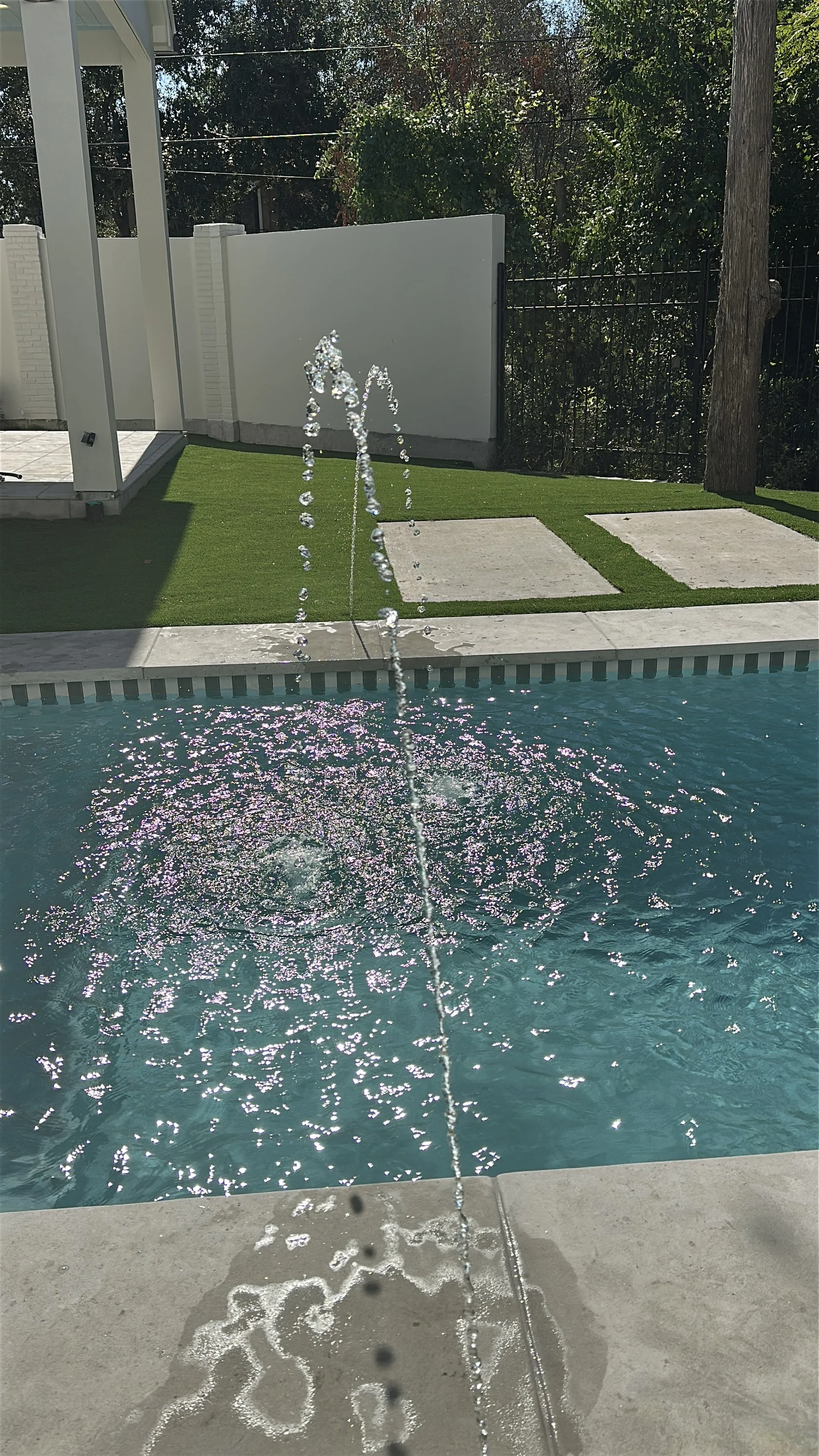 A swimming pool with a fountain feature and a patch of artificial grass next to a white privacy wall and a black metal fence in a backyard.