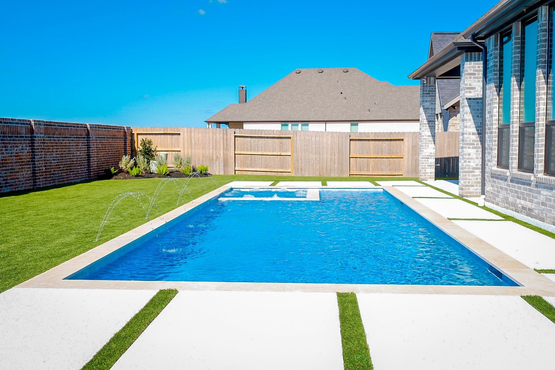 Backyard with a rectangular swimming pool, green grass, a wooden fence, and a brick house under a clear blue sky.
