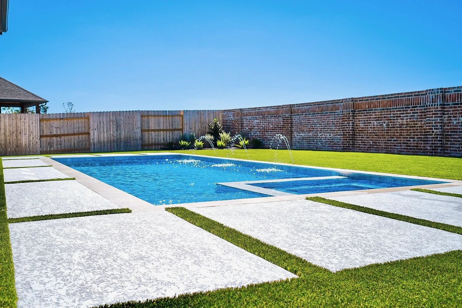 Backyard with a swimming pool, white concrete stepping stones, green grass, a wooden fence, a brick wall, and blue sky.