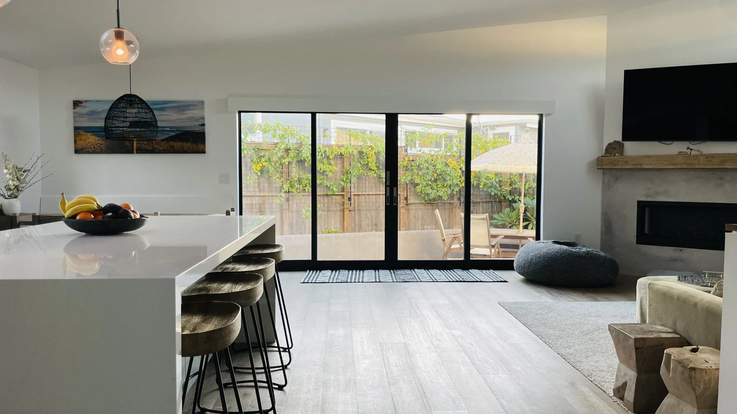 Modern living room with large sliding glass doors opening to a patio with outdoor seating, a plant, and a garden. The room features a white kitchen island with a bowl of bananas and fruit, a wall-mounted TV above a fireplace, a large gray bean bag, a