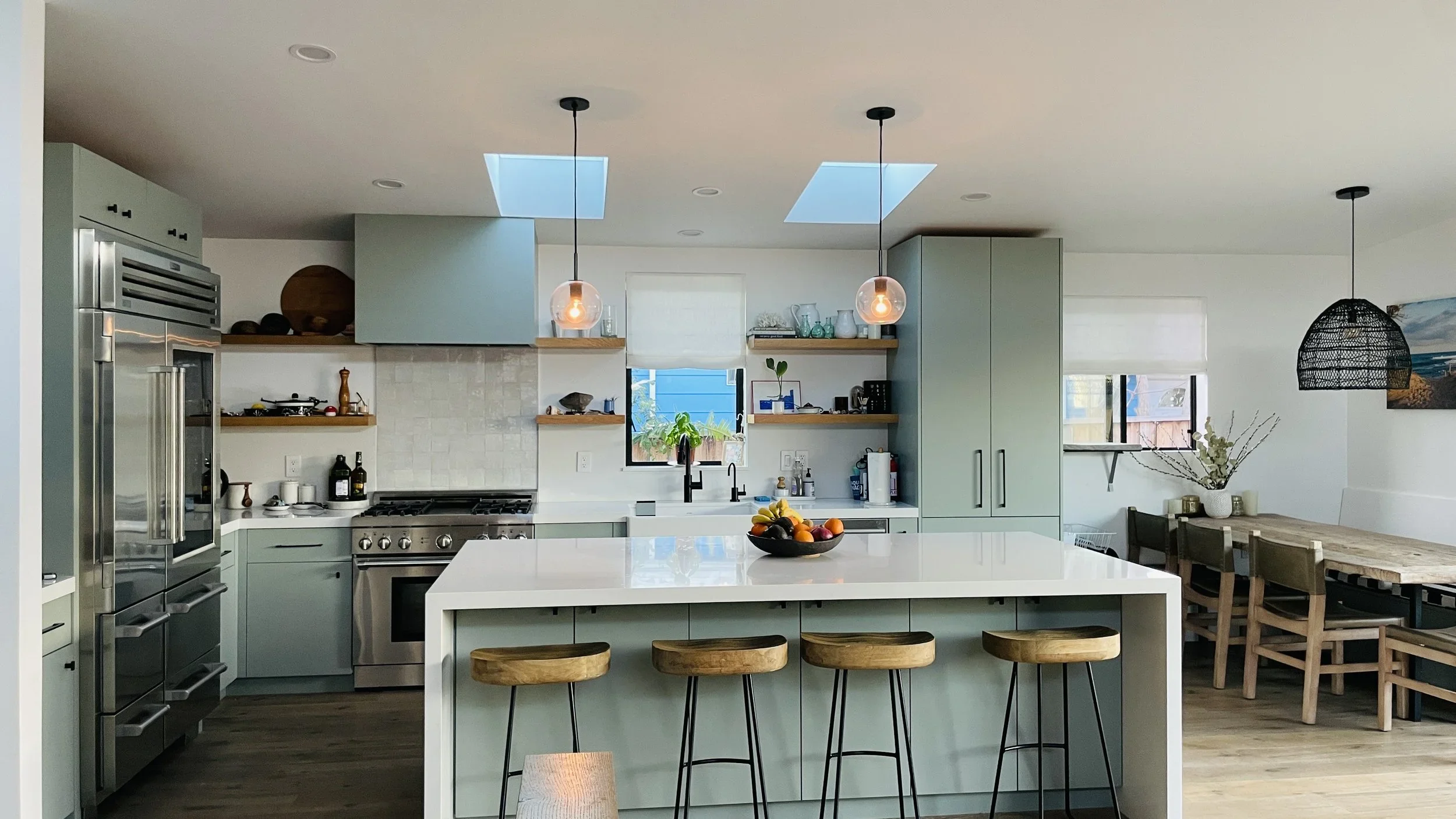 Modern kitchen with white island, wooden stools, stainless steel appliances, open shelving, and a dining table with chairs in the background.