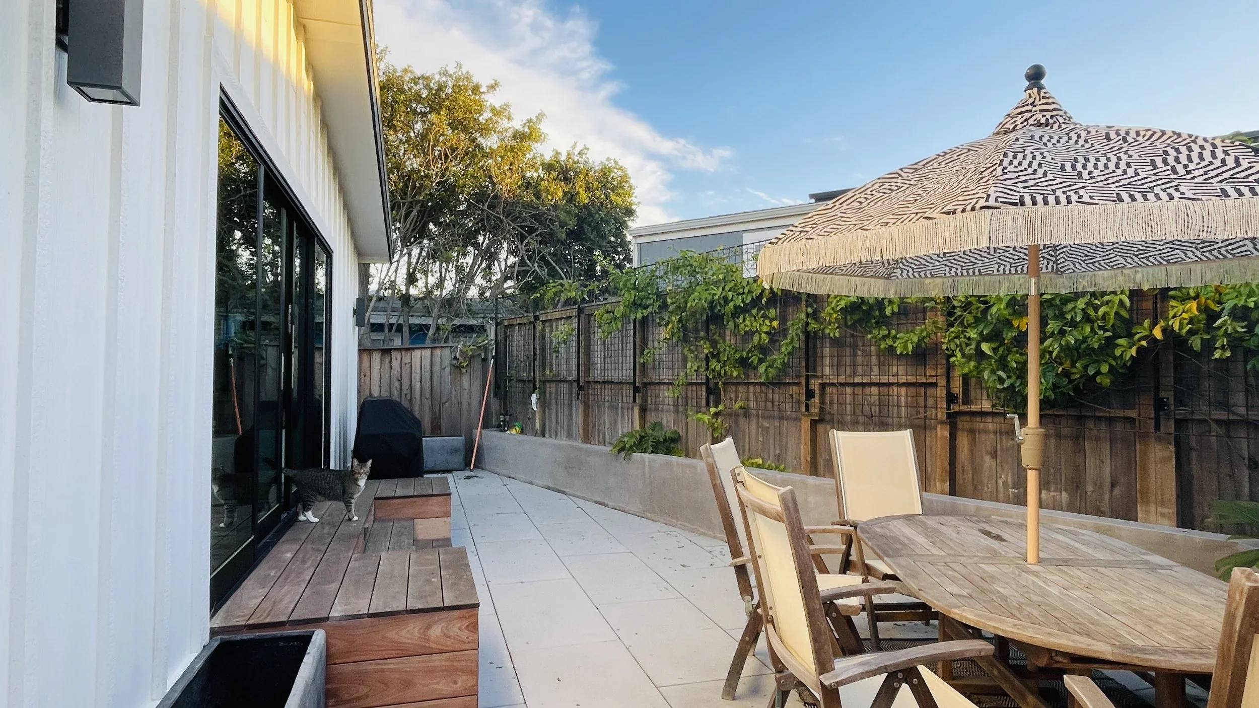 A backyard patio with a wooden table and chairs, a large umbrella, a cat near sliding glass doors, and a fenced garden with trees and plants, under a partly cloudy sky.