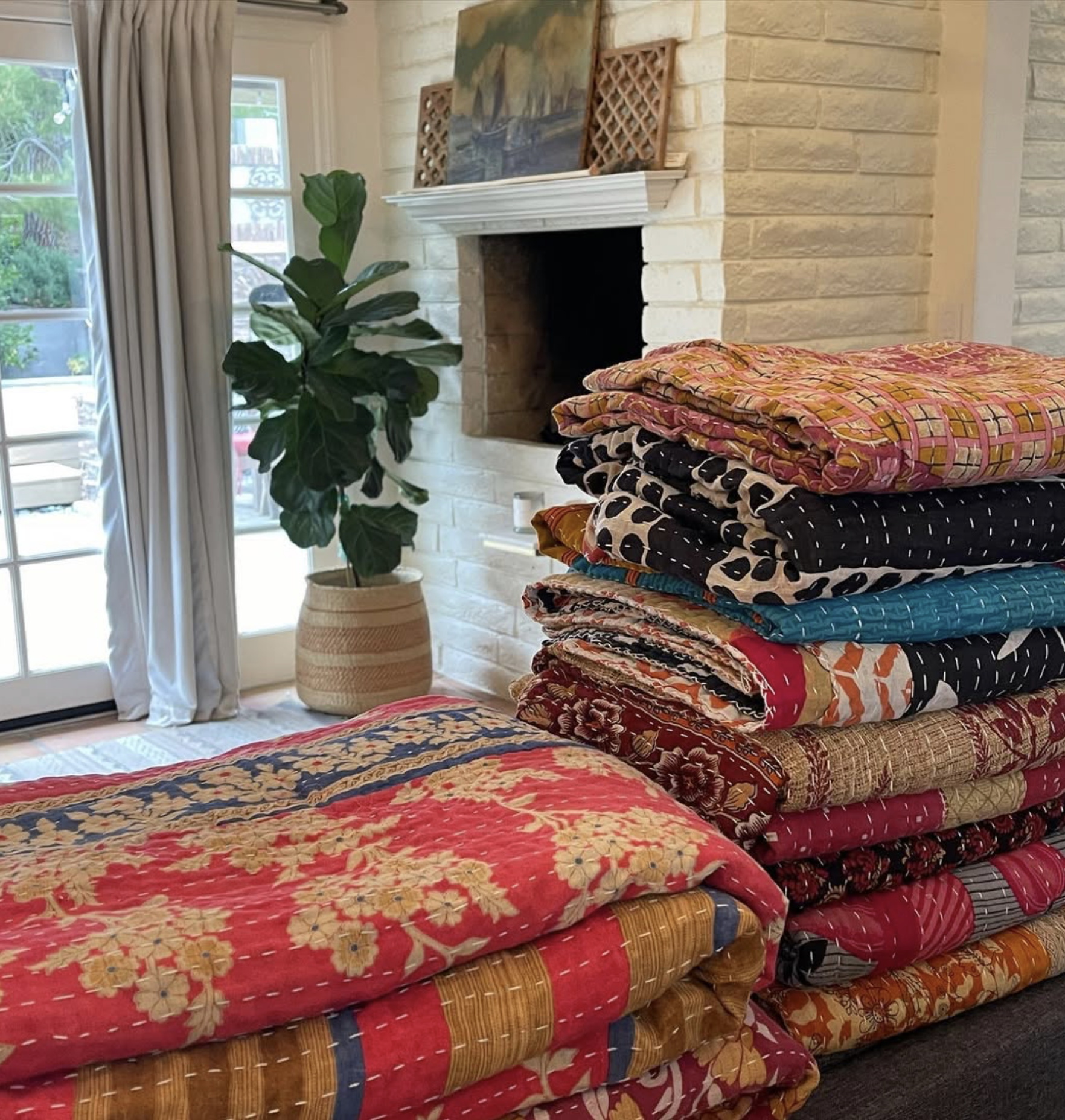 Stacked colorful quilts and a quilted blanket on a dark piece of furniture in a cozy living room with a white brick fireplace, a large green potted plant, and a glass door with curtains.