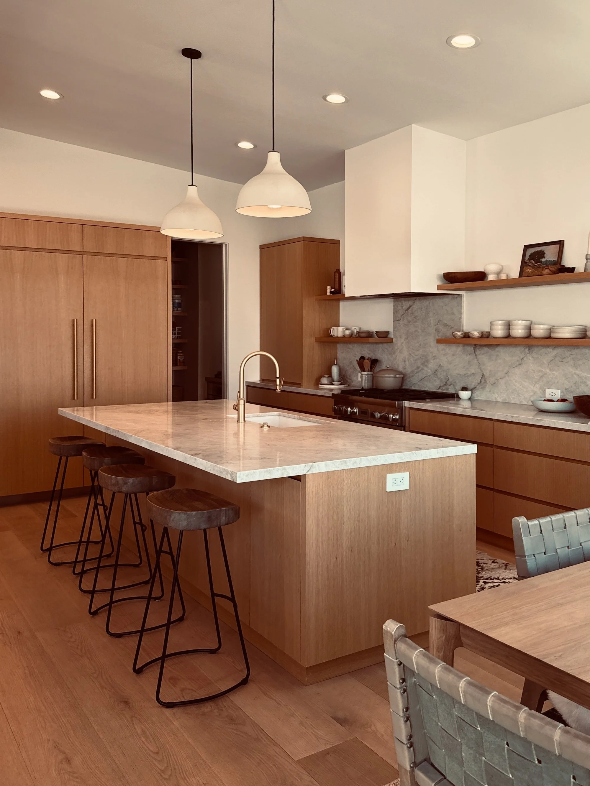 Modern kitchen with wooden cabinets, a marble island with a sink, and open wooden shelves displaying dishes and decorative items. Two pendant lights hang above the island, and a dining table with woven chairs is visible.