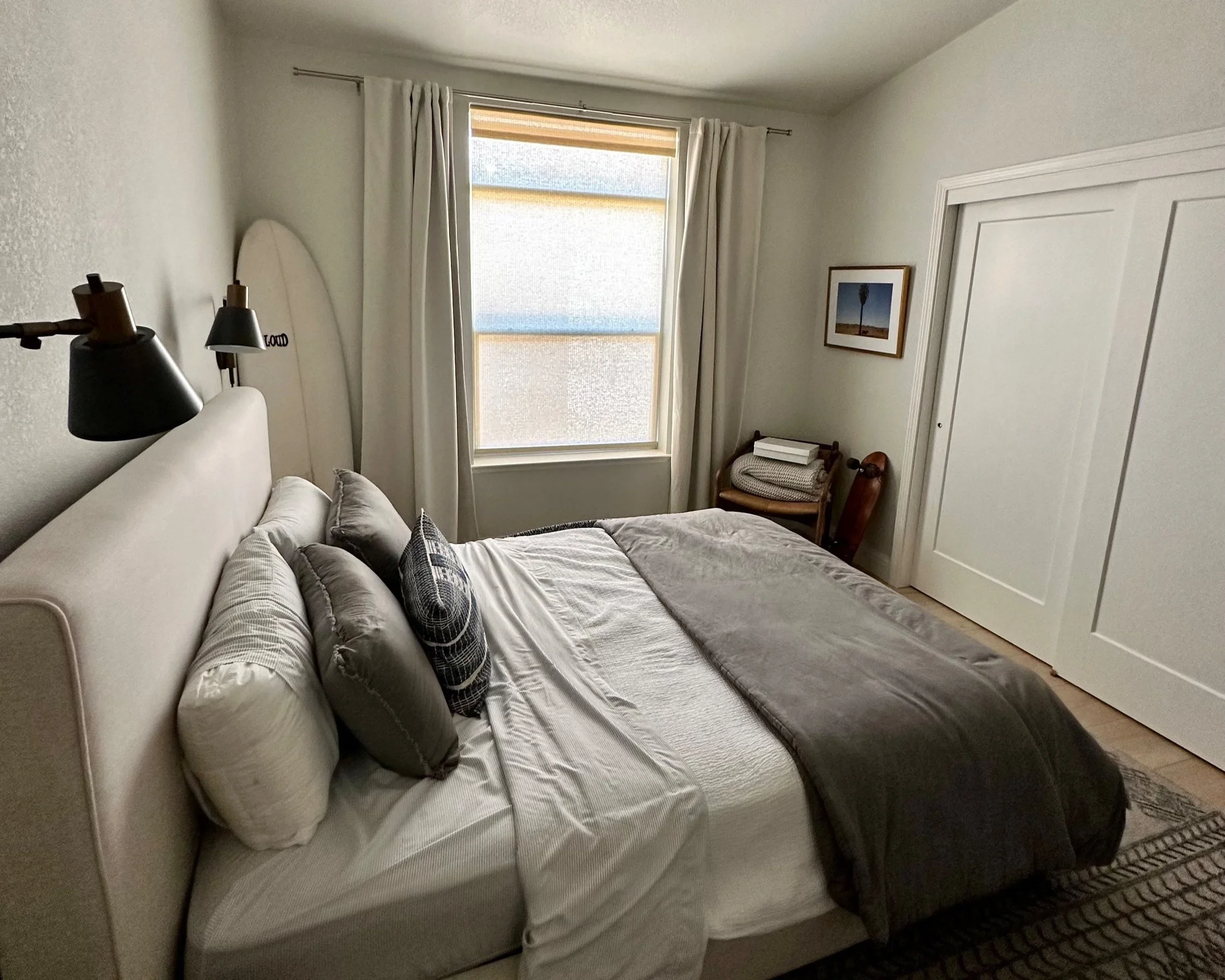 A neatly made bed with white and gray bedding, decorative pillows, and black wall-mounted lamps in a minimalist bedroom with a window, framed artwork, and a chair in the corner.