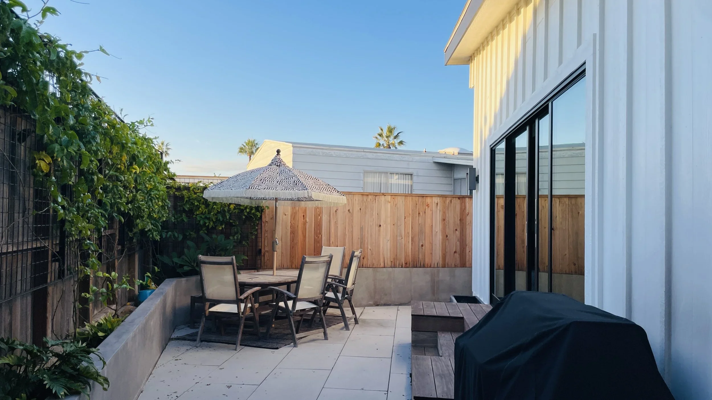 Residential backyard patio with outdoor dining table, chairs, umbrella, greenery, wooden fence, sliding glass door, and barbecue grill under a clear blue sky.