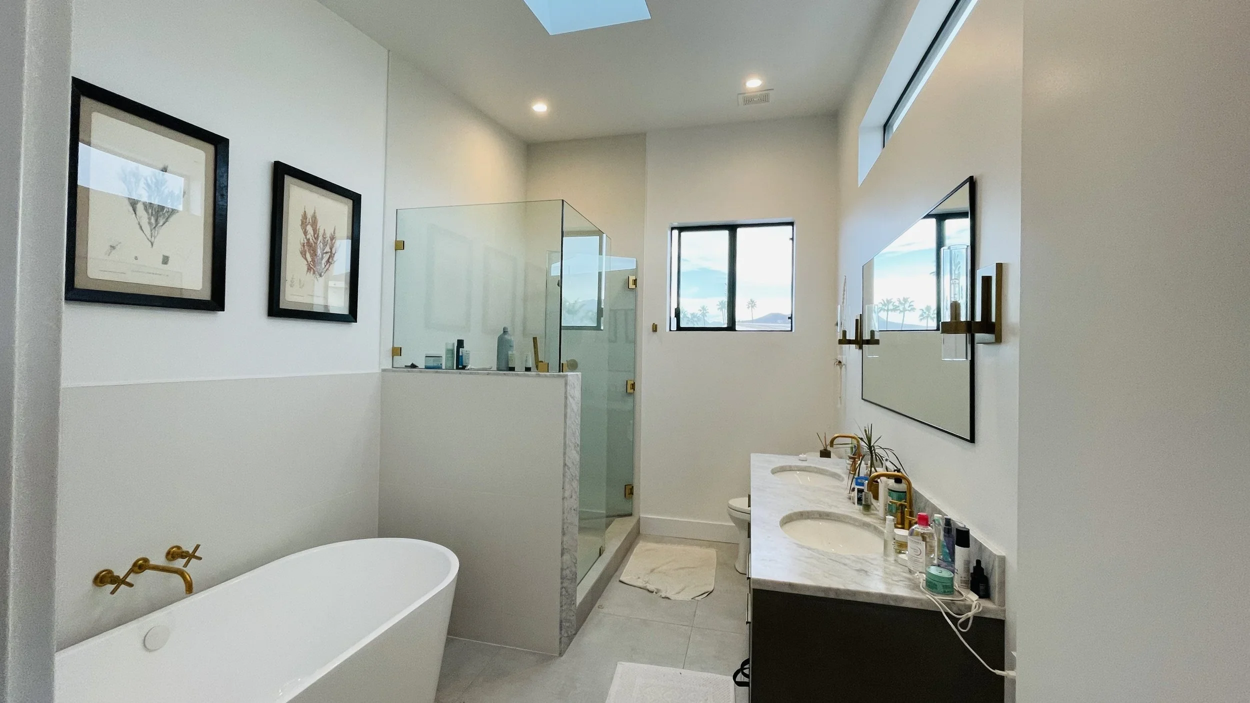 Modern bathroom with a freestanding bathtub, glass-enclosed shower, dual sinks with marble countertop, large mirror, and framed botanical art on white walls, with windows showing palm trees outside.