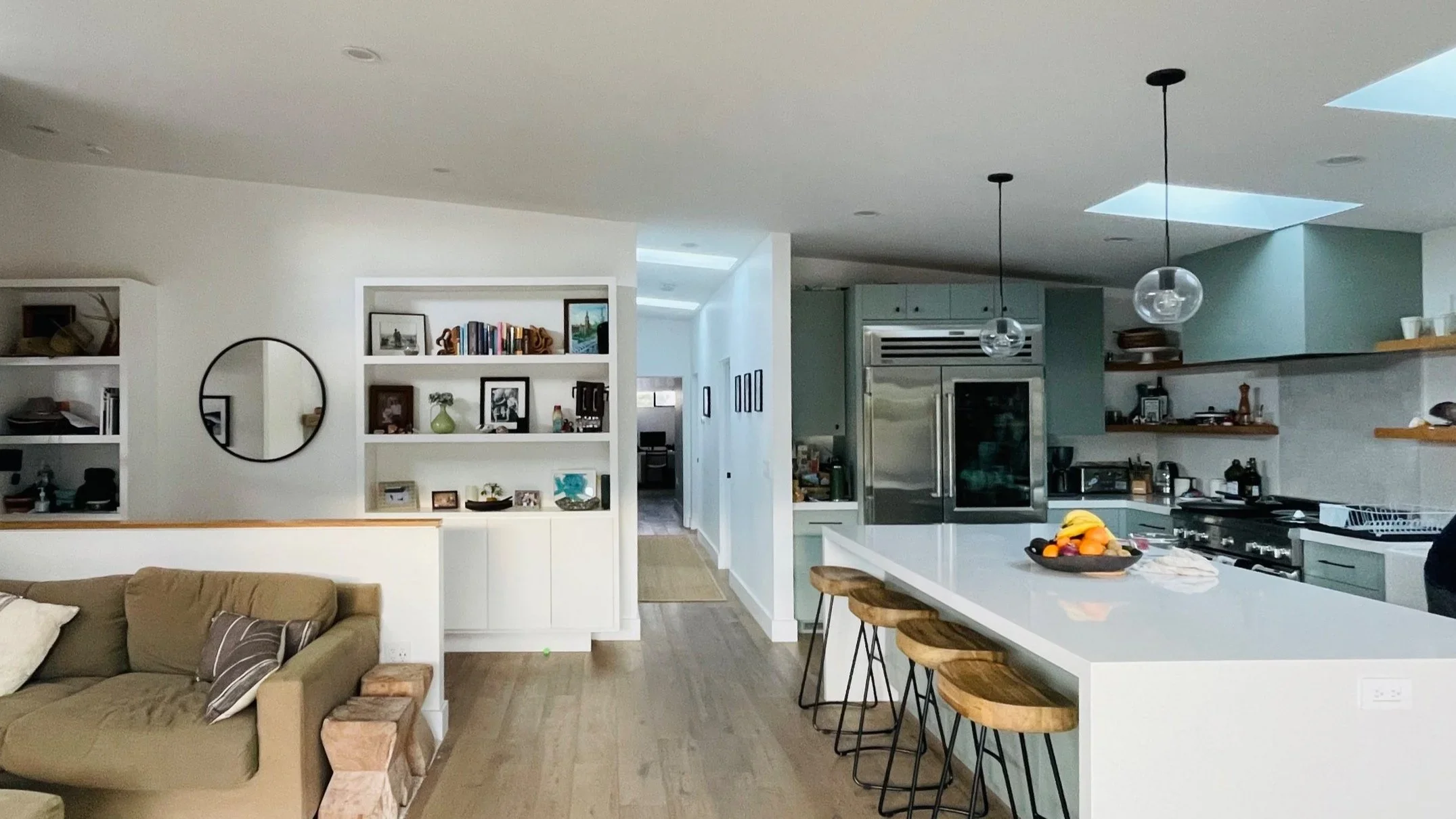 Modern open-concept kitchen with a large white island, wooden bar stools, stainless steel appliances, and a cozy living area with a beige sofa and decorative shelves. Skylights in the ceiling provide natural light.