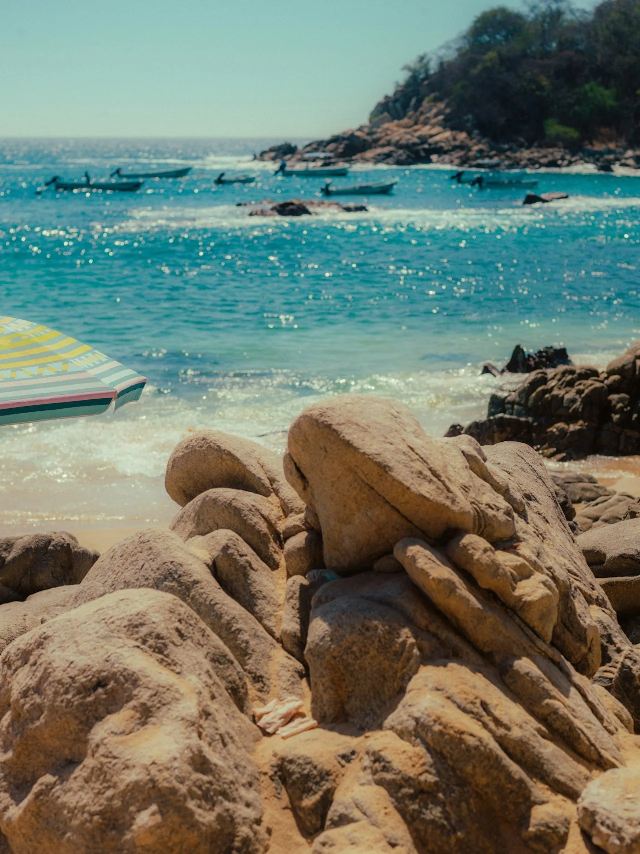 Rocky beach with large rocks in the foreground, ocean waves, several boats, and a hillside covered with trees in the background.