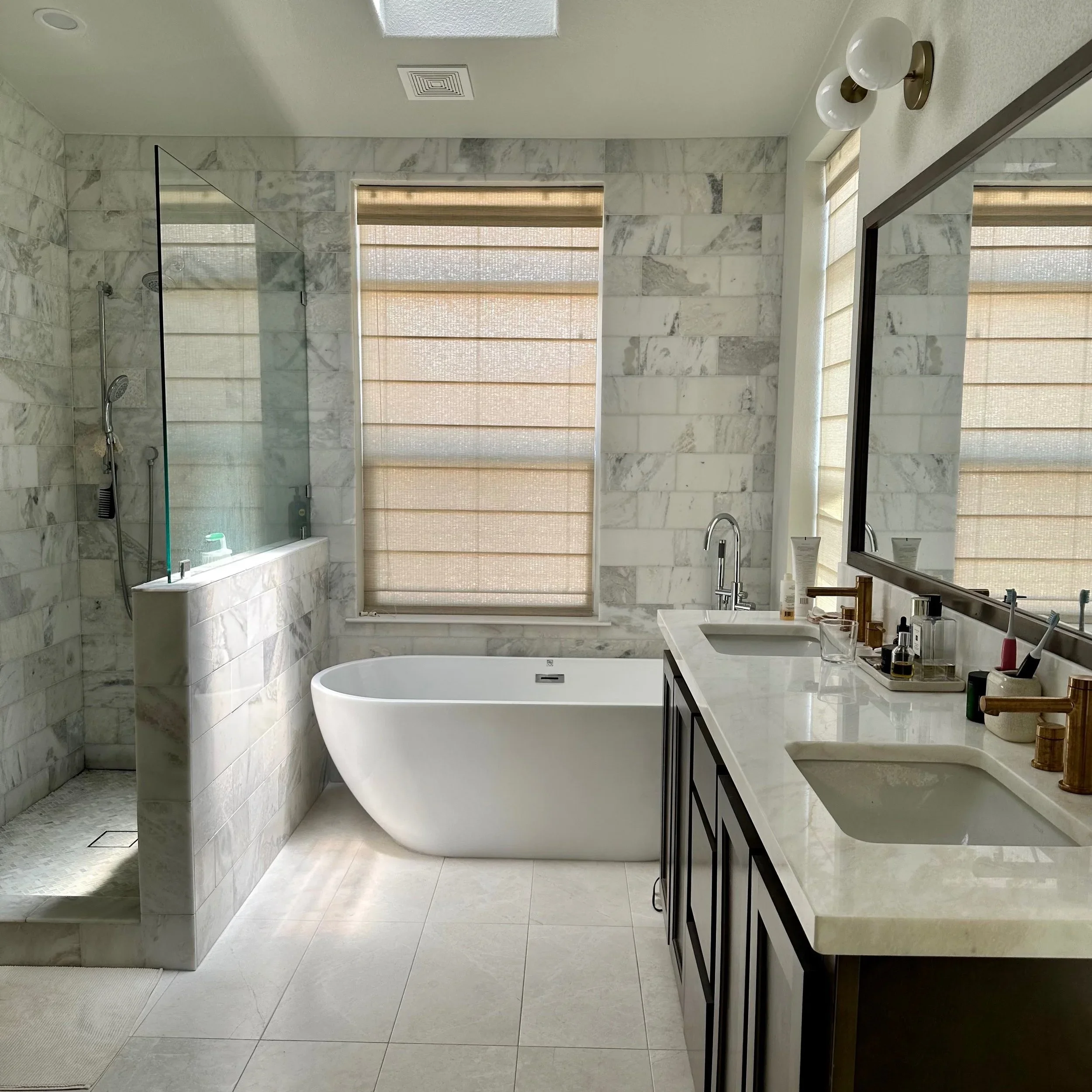 Modern bathroom with marble walls, a freestanding bathtub, a large mirror, and a double sink vanity with various toiletries. There is a walk-in shower with a glass partition and a window with beige blinds.