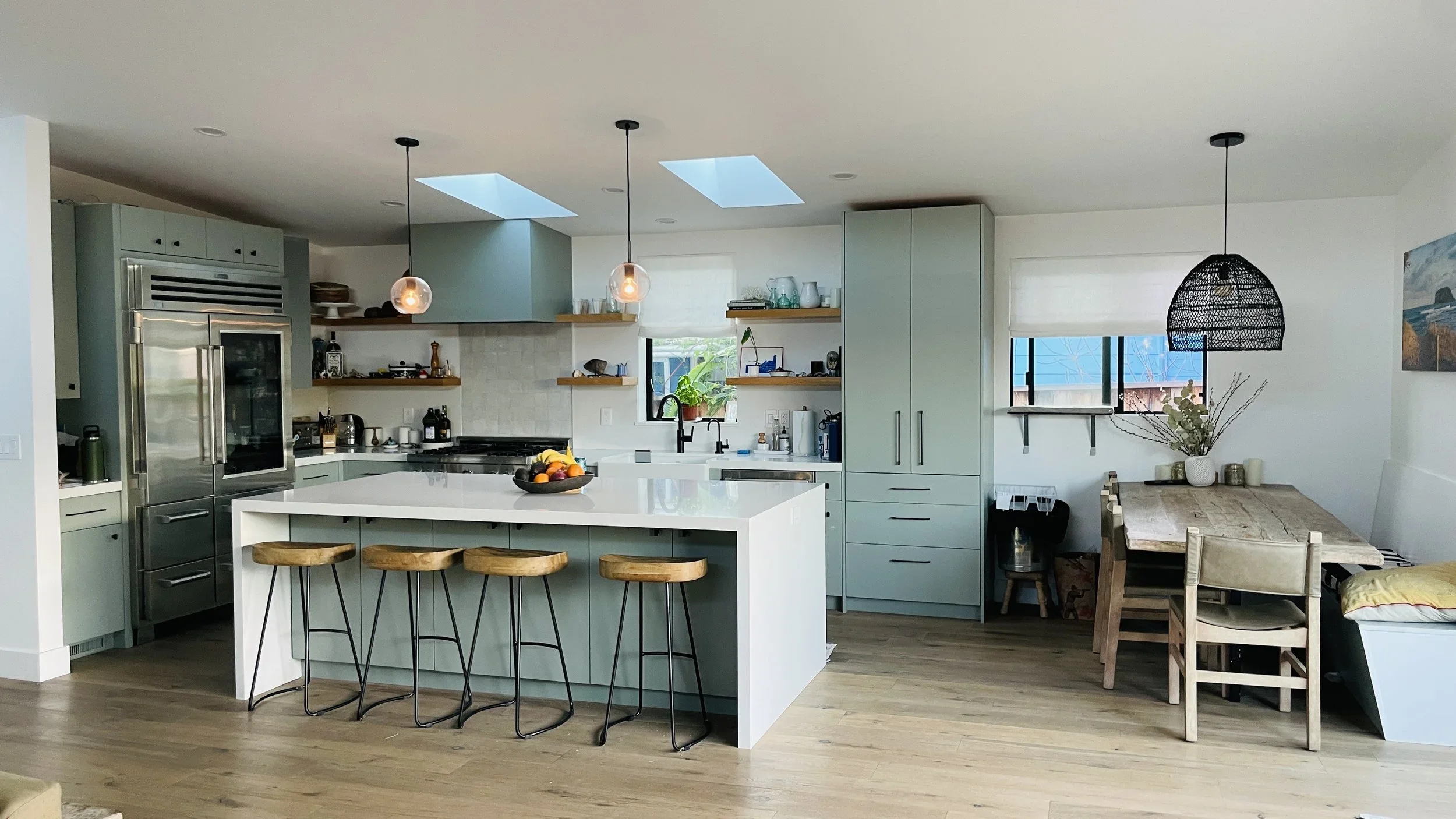 Modern kitchen with gray cabinets, white island with four wooden stools, stainless steel appliances, open shelving, black pendant lights, and a dining area with a wooden table and chairs near a window.