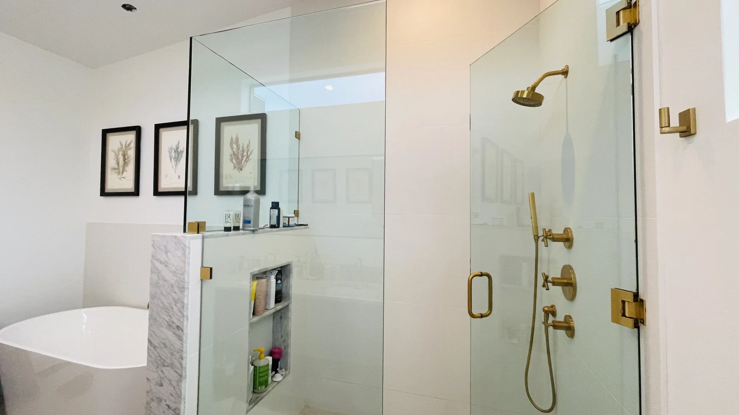 A modern bathroom with a glass shower enclosure featuring a gold showerhead and fixtures, a white bathtub with a marble accent, and three framed botanical prints on the wall.