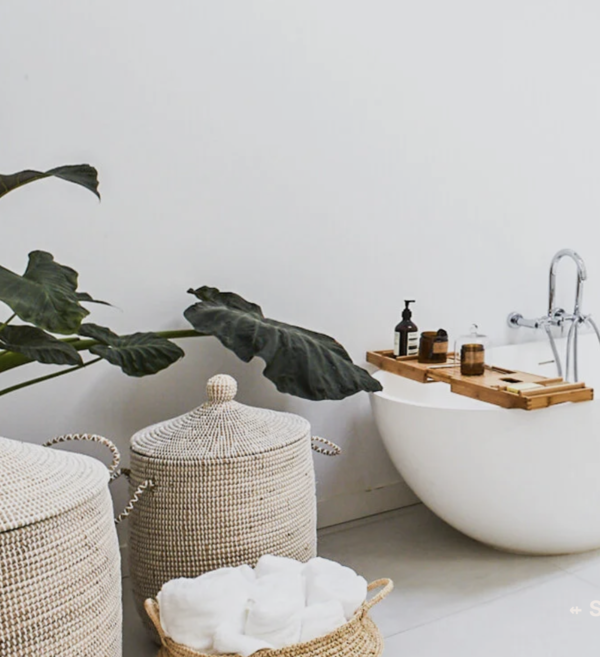 A minimalist bathroom corner with a white bathtub on the floor, a wooden bath tray holding bottles, a large green plant with broad leaves, and woven baskets for storage.
