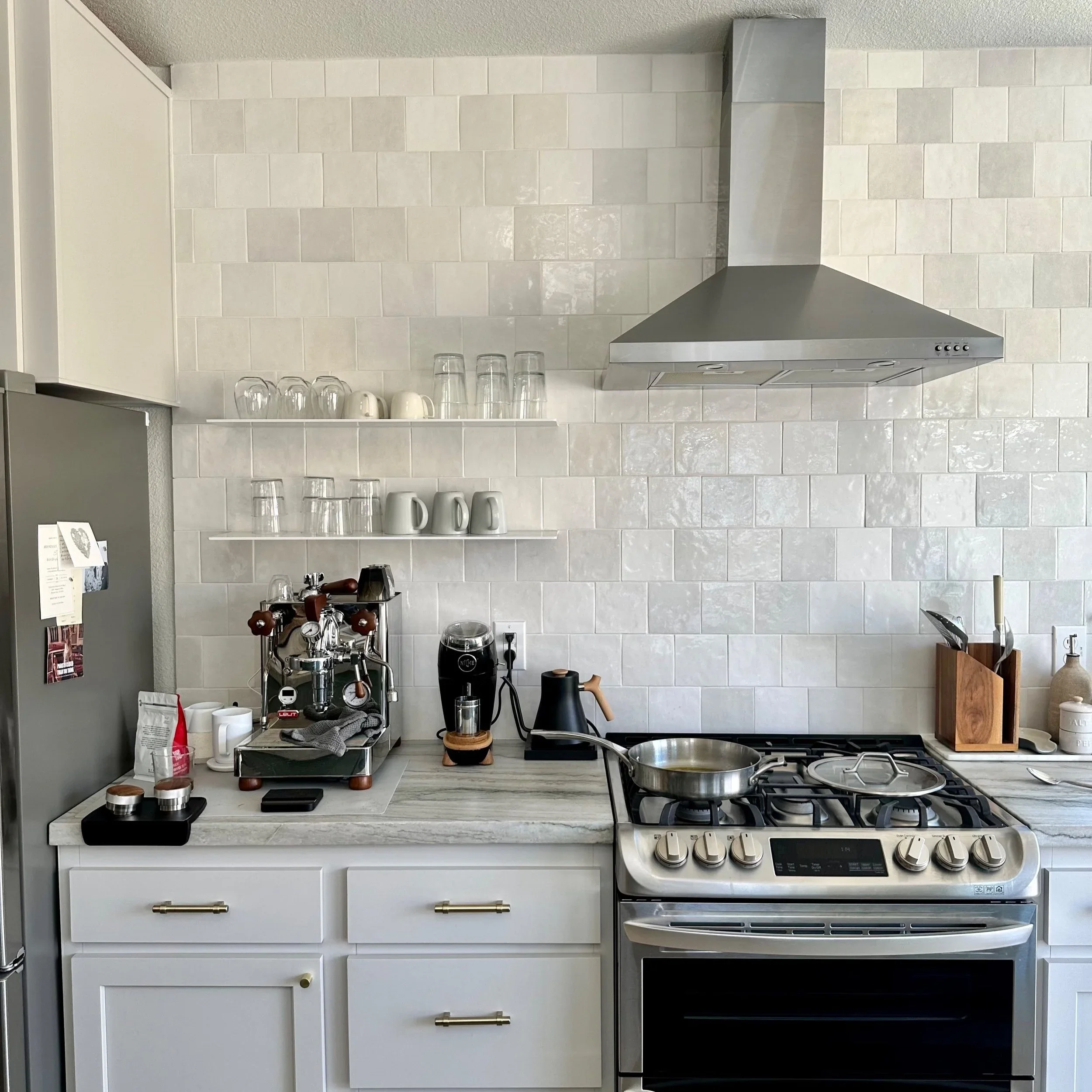Modern kitchen with white cabinets, gray countertop, wall-mounted shelves with glassware and mugs, stainless steel range hood, and stainless steel gas stove with an empty frying pan.