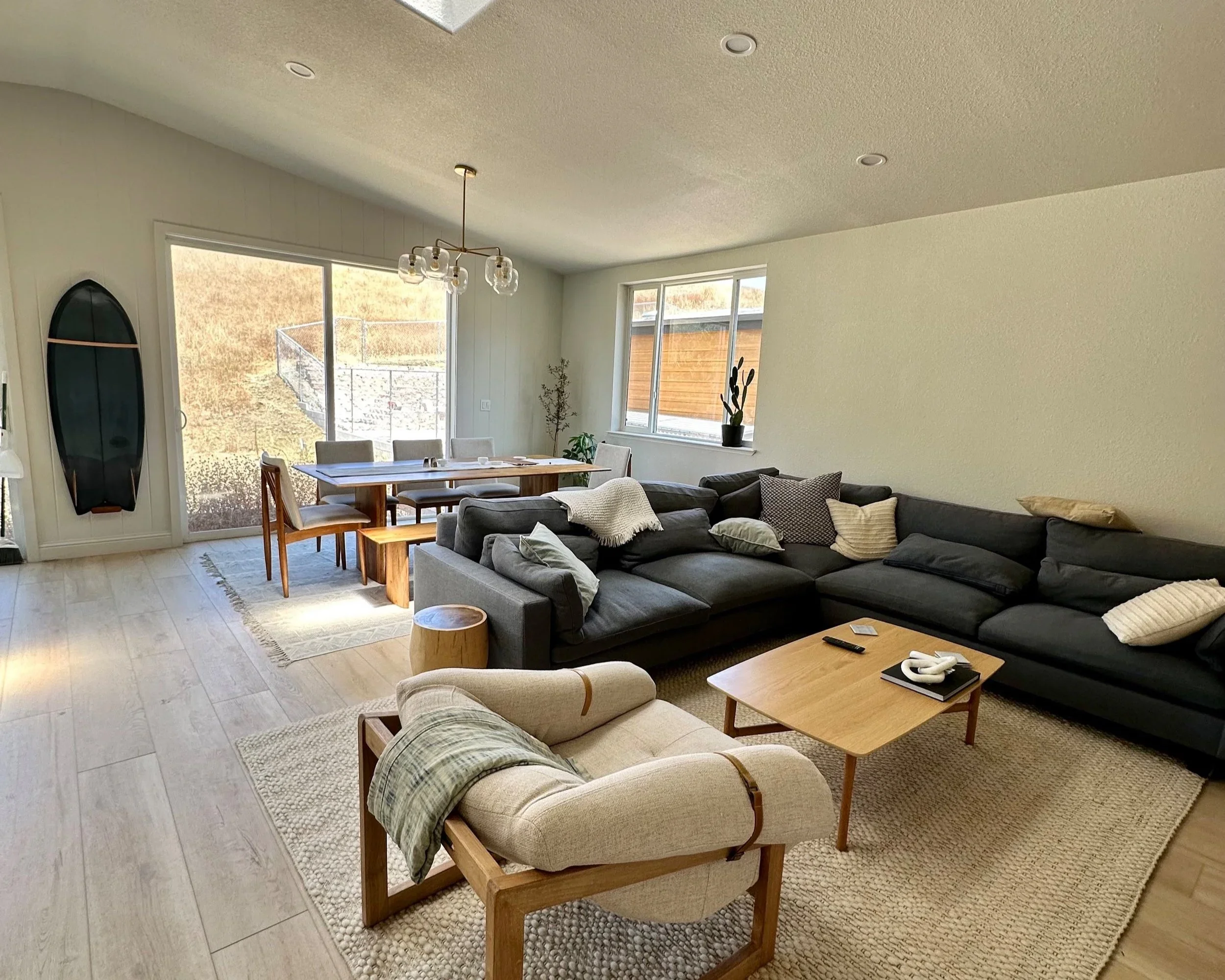 Living room with beige armchair, dark gray sectional sofa with pillows, wooden coffee table, and dining area with wooden table and chairs, large windows, and modern chandelier.