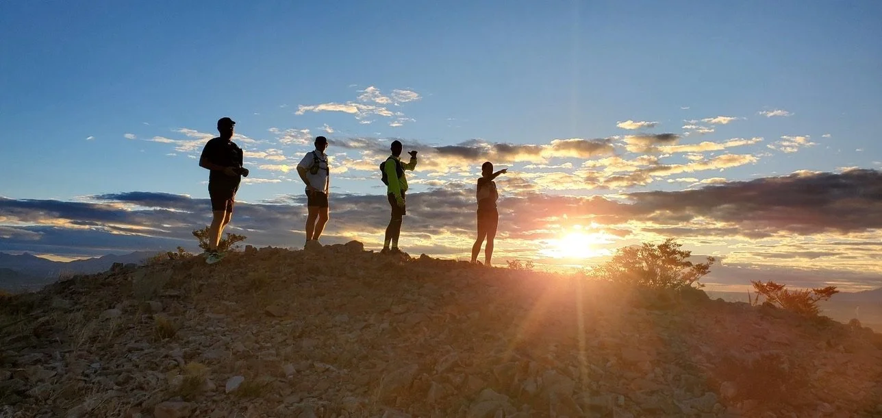 Four hikers standing on a mountain ridge with the sun low on the horizon.
