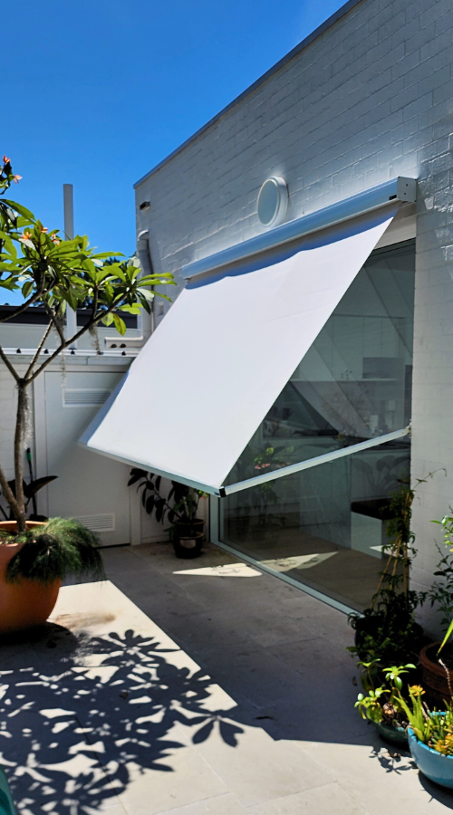 A white awning extending from the side of a building, shading a glass door and patio area with potted plants and trees, under a clear blue sky.