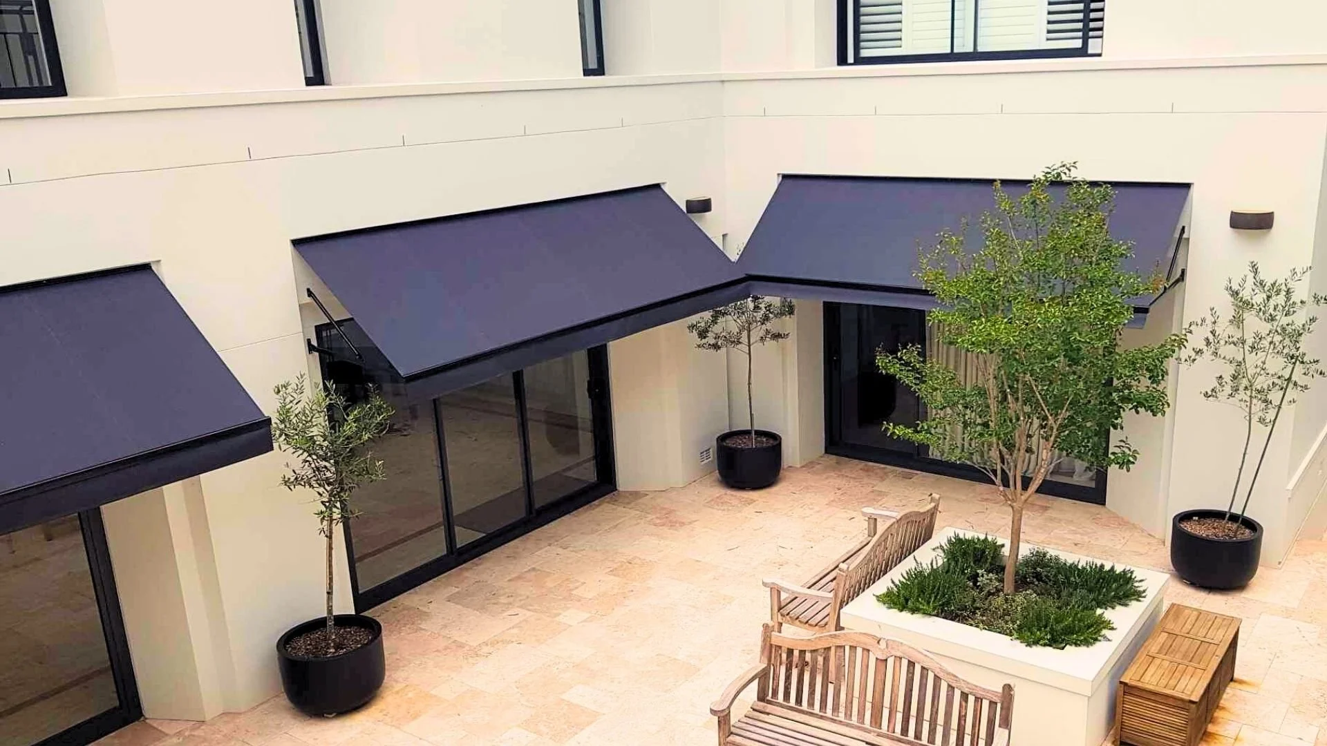 Interior courtyard with beige tiled floor, two trees in black planters, a large planter with a tree and greenery, two wooden benches, and black awnings on the building's exterior walls.