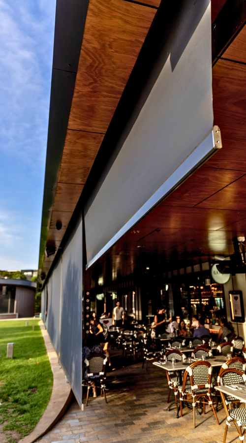 Outdoor patio area of a restaurant or cafe with tables and chairs, and people dining inside. The building has a wooden ceiling and a large retractable outdoor shutter.