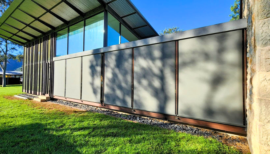 Modern building exterior with large glass windows, metal panels, and a sloped roof, surrounded by a grassy lawn and shadows of trees.