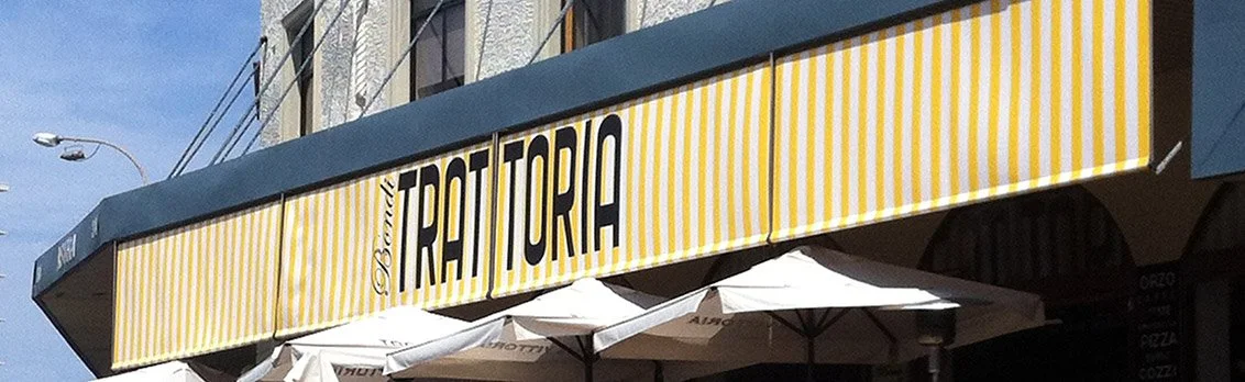 Yellow and white striped awning with the words 'Restaurante Trattoria' in bold black letters, white umbrellas outside the restaurant, and part of a building with windows.