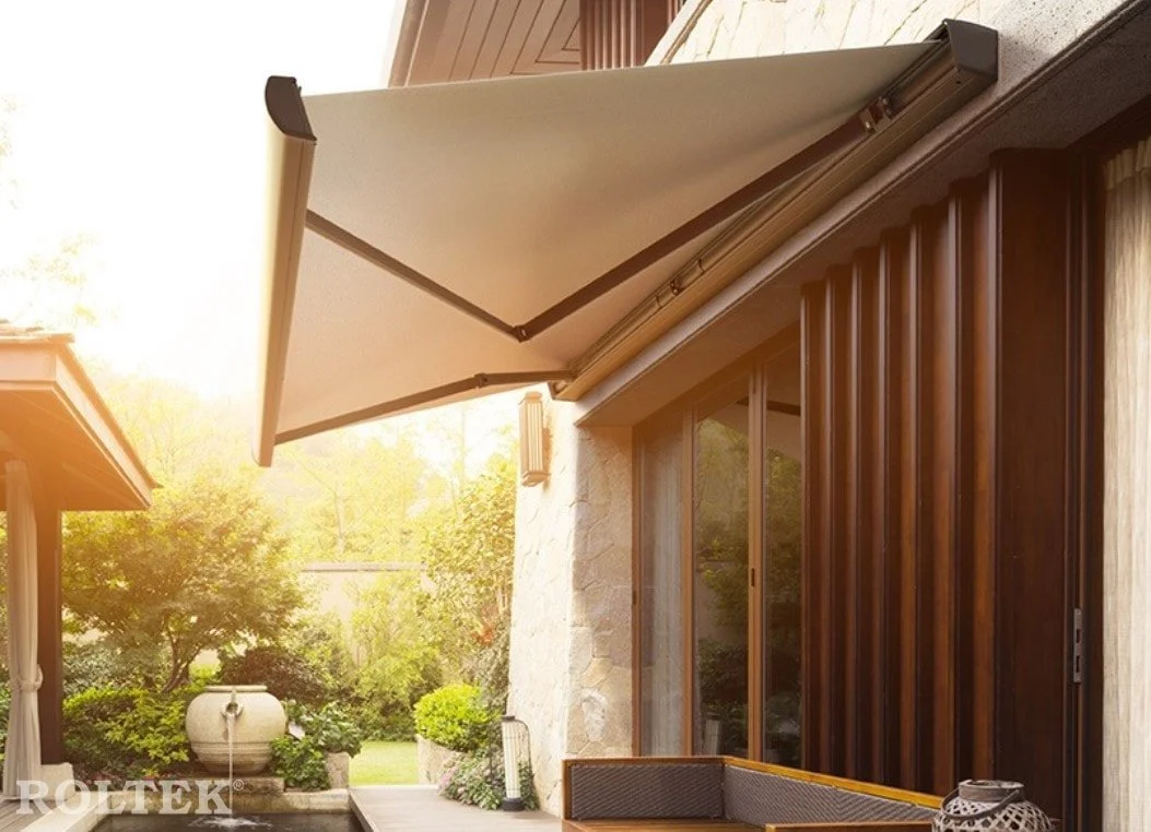A patio area with a beige retractable awning, wooden siding on the house, glass doors, and outdoor furniture in a garden setting with trees, bushes, and a large ceramic planter.