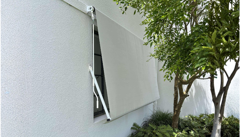 A window with a white retractable sunshade on a white wall, next to a green shrub and tree.