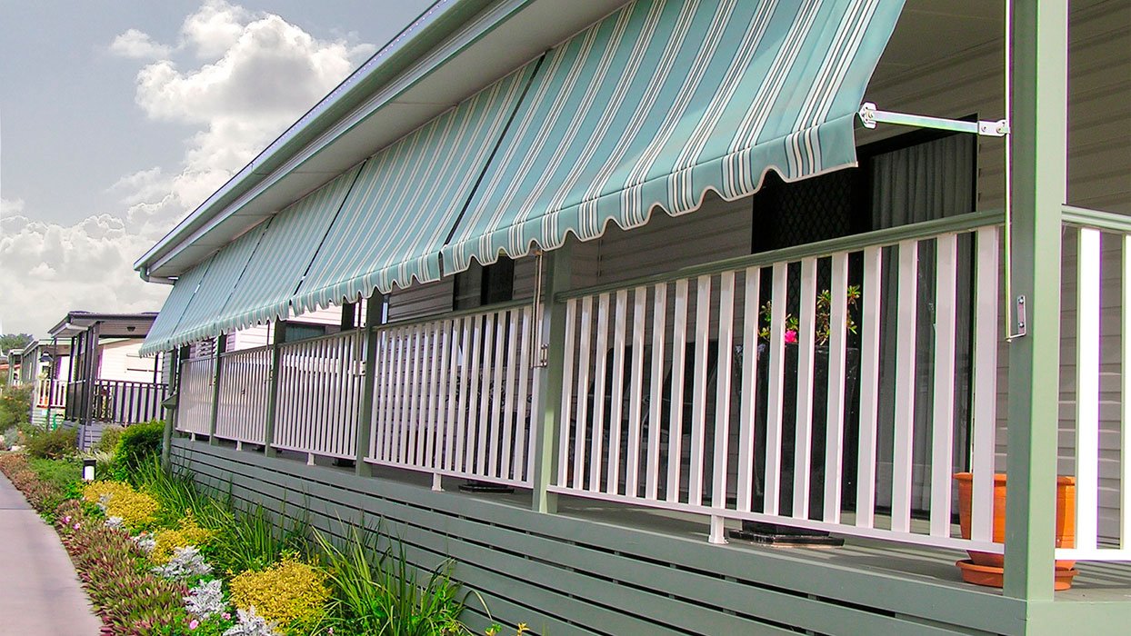 A residential house with a porch, white railing, and a green-and-white striped awning. There are potted plants and flowers on the porch, and a sidewalk with colorful flowers along the house.