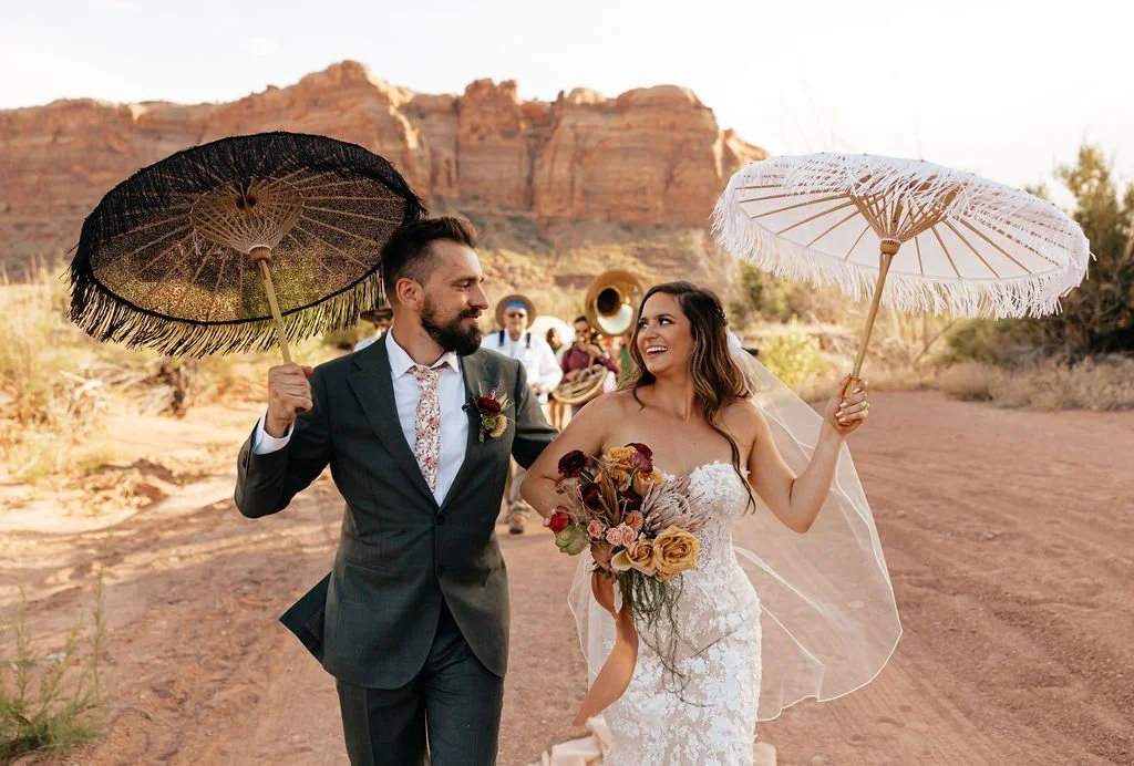 A Groom and Bride With Umbrellas Overhead Lead Their Wedding Guest In A New Orleans Traditional Second Line March Below Sandstone Cliffs at The Red Earth Venue Moab, Utah
