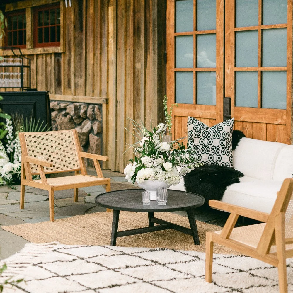 A Modern Styled Lounge With Natural Wood and Rattan Chairs, A White and Black Tassel Rug, Black Coffee Table, White Rose Center Piece, And a White Couch