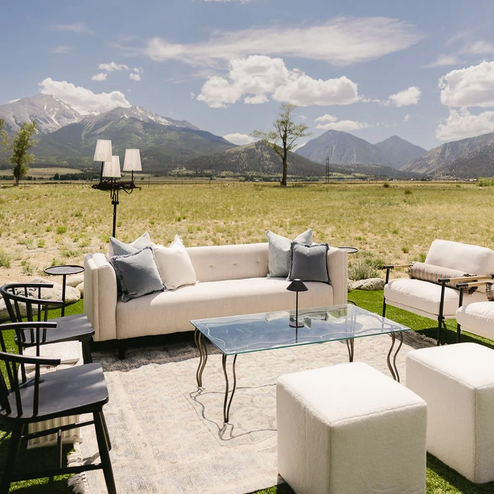 A Wedding Reception Lounge Setup In a Meadow Near Colorado Rocky Mountains. A White Couch, White Ottoman, Glass Coffee Table, and Black Wood Chairs Make Up This Lounge.