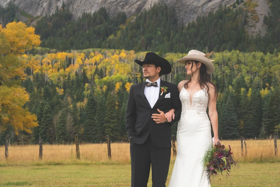 A Groom Dressed In A Formal Tuxedo With A Cowboy Hat Stands Arm in Arm With His Bride Dressed In A Modern Lace Dress With A Beige Cowboy Hat With Large Granite Cliffs Behind Them At The Sombrilla Springs Wedding Venue
