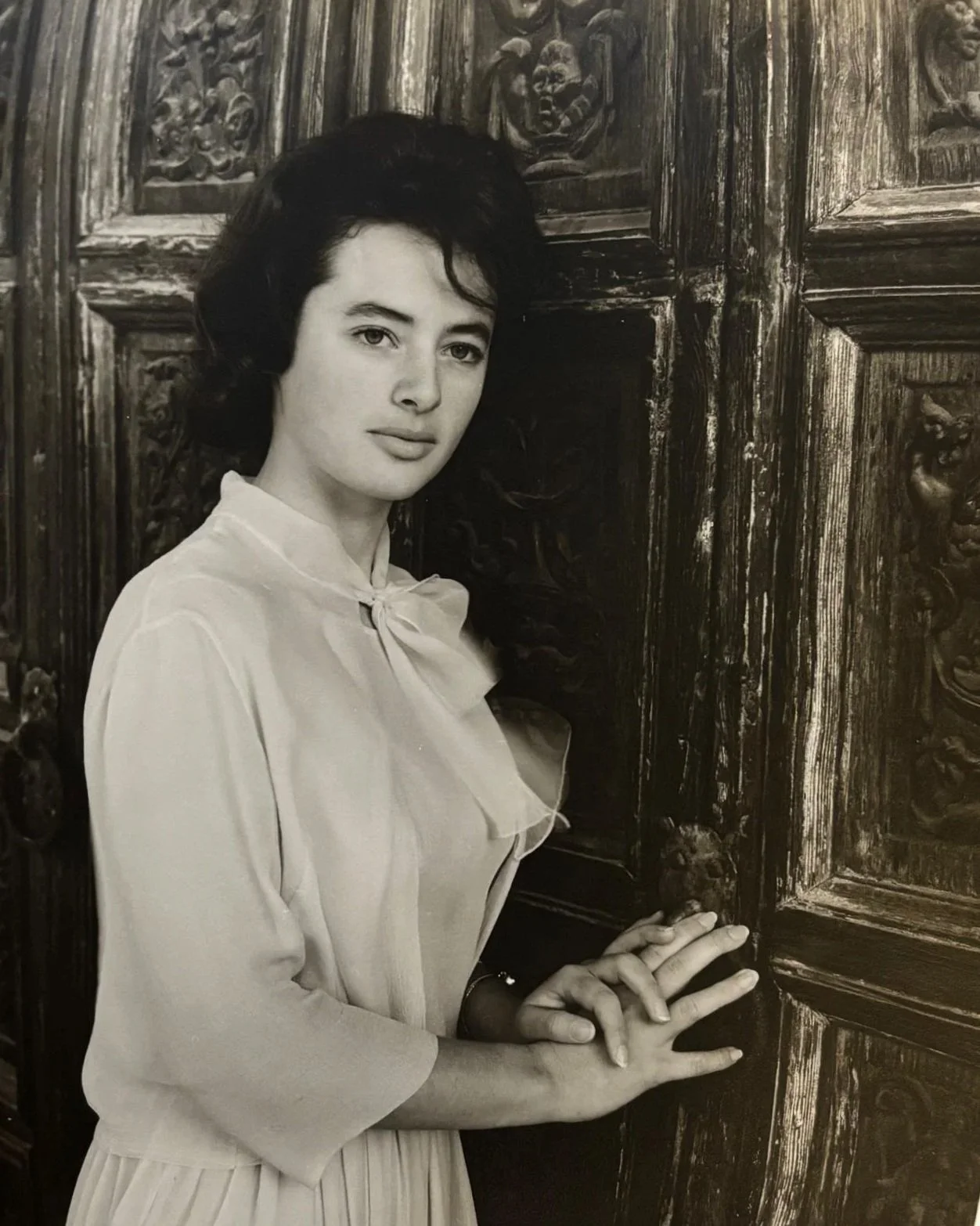 Black and white portrait of a woman with dark hair, wearing a light blouse with a bow, standing against ornately carved wooden panels.