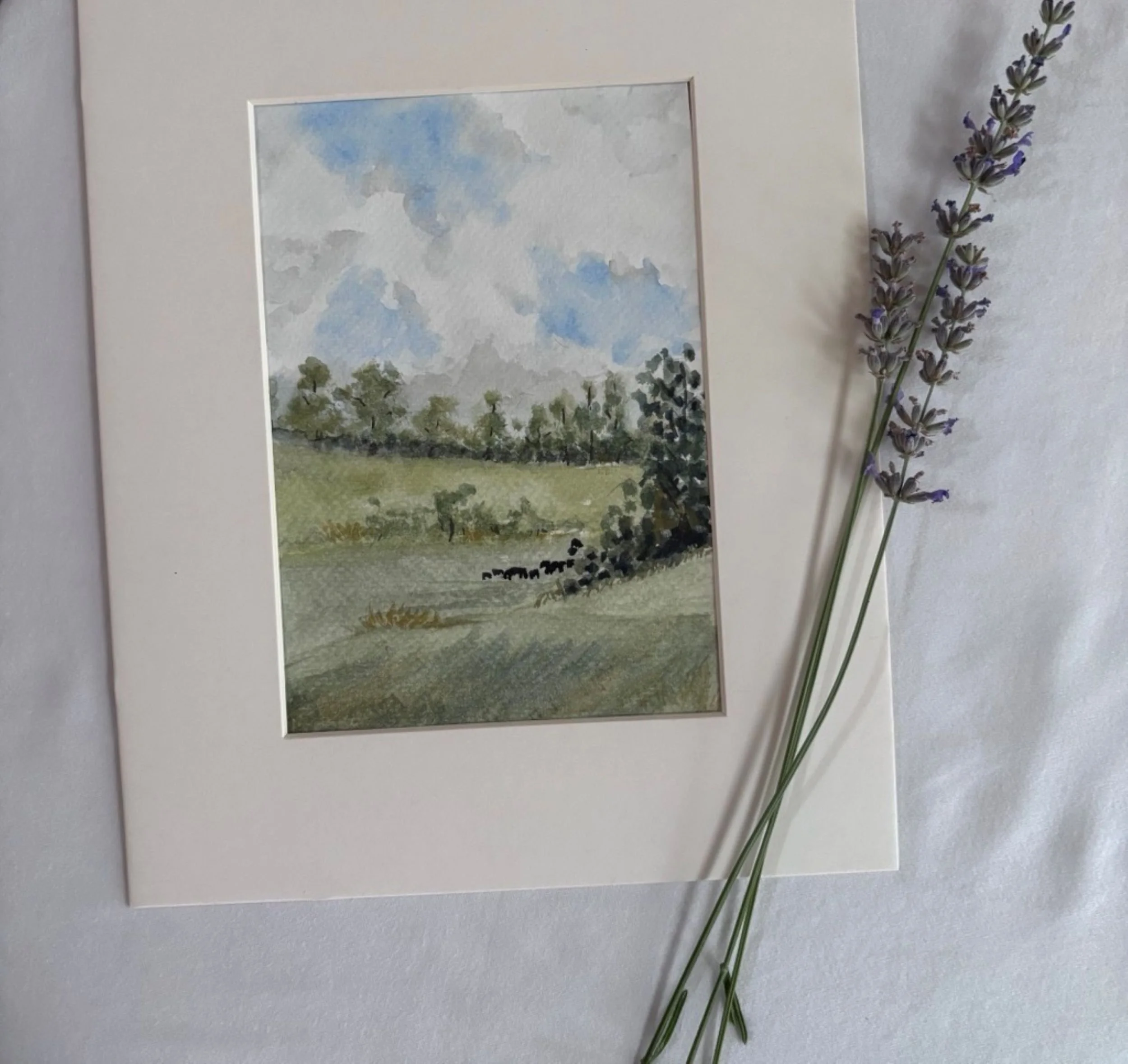 A watercolor painting of a landscape with trees under a cloudy sky, resting on a white surface with a sprig of lavender flowers beside it.
