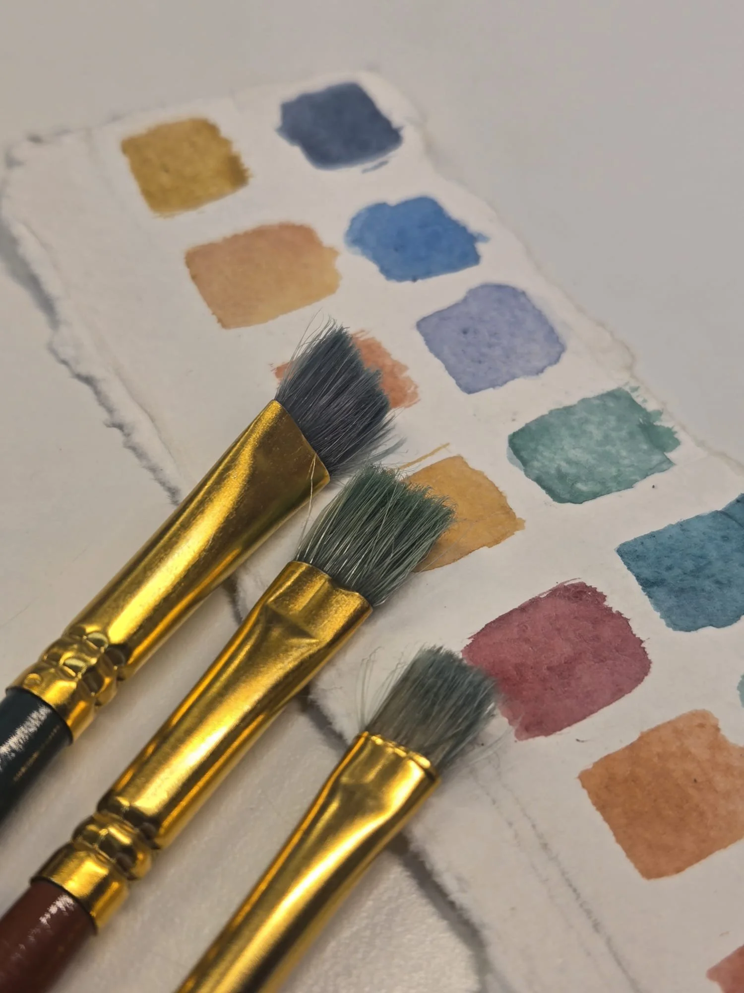 Close-up of watercolor paint swatches in various colors with paintbrushes on a textured paper.