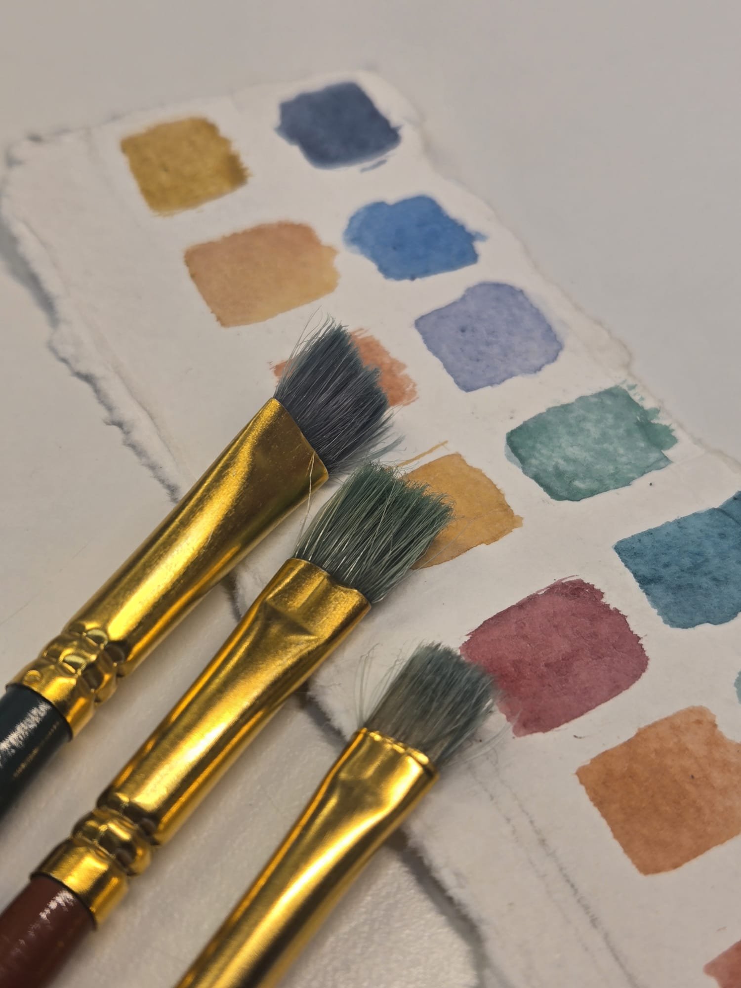 Close-up of watercolor paint swatches in various colors with paintbrushes on a textured paper. Watercolor painting supplies, brushes, and artist color palette used to create original watercolor artwork.