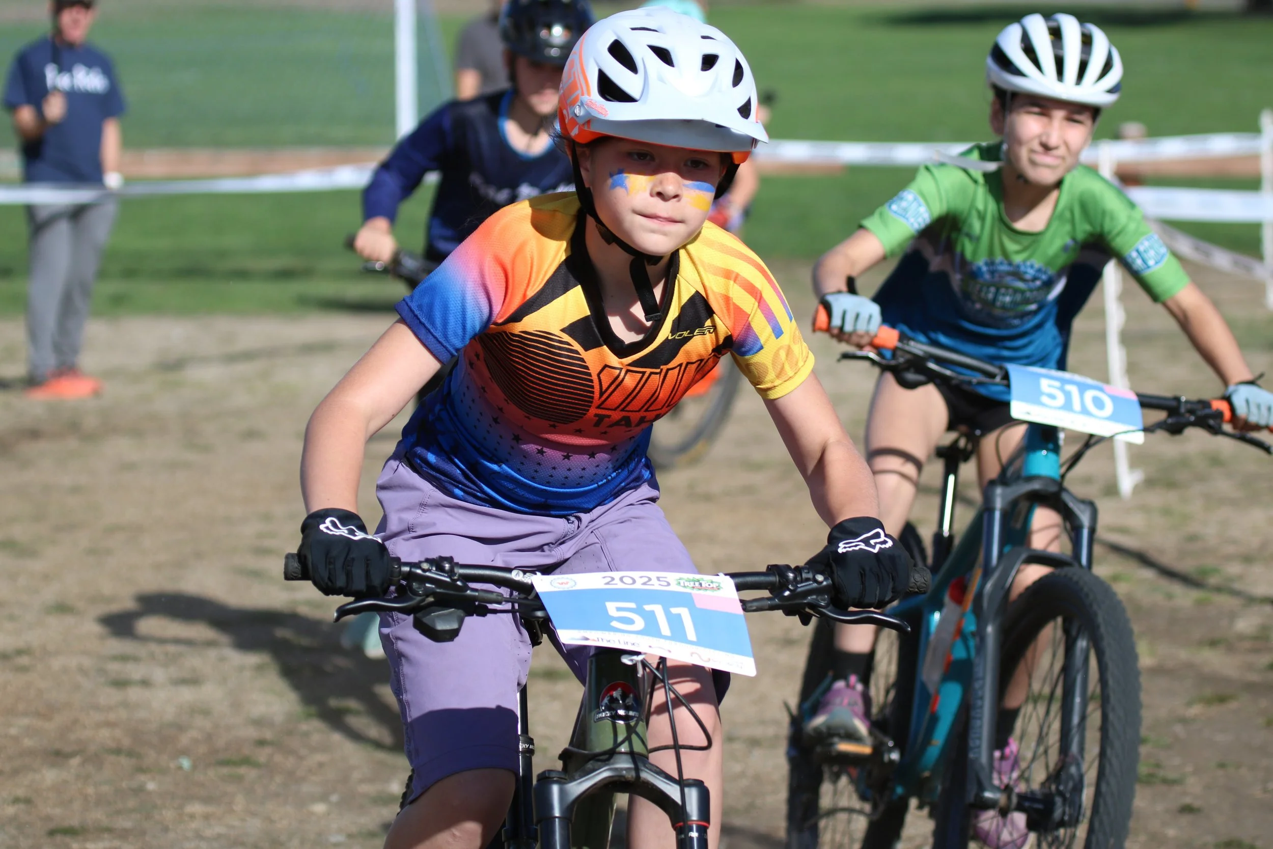 Children participating in a bike race, wearing helmets and race numbers, outdoors on a dirt track.