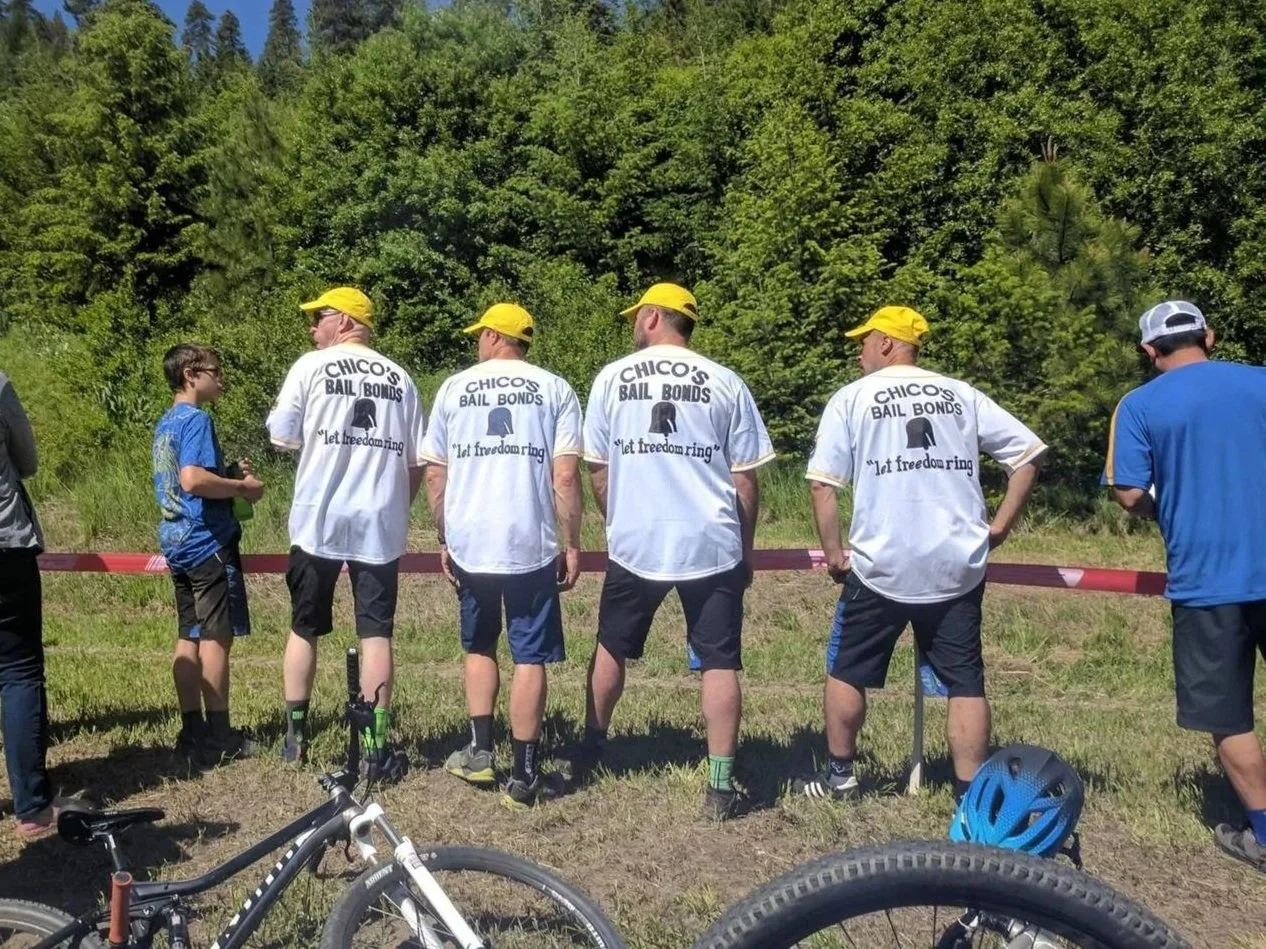 Group of people standing outdoors next to a red rope, with lush green trees in the background. Several individuals are wearing white shirts with 'Chico's Bail Bonds' and the slogan 'let freedom ring,' some are wearing yellow caps. Bicycles and a blue helmet are on the ground in the foreground.