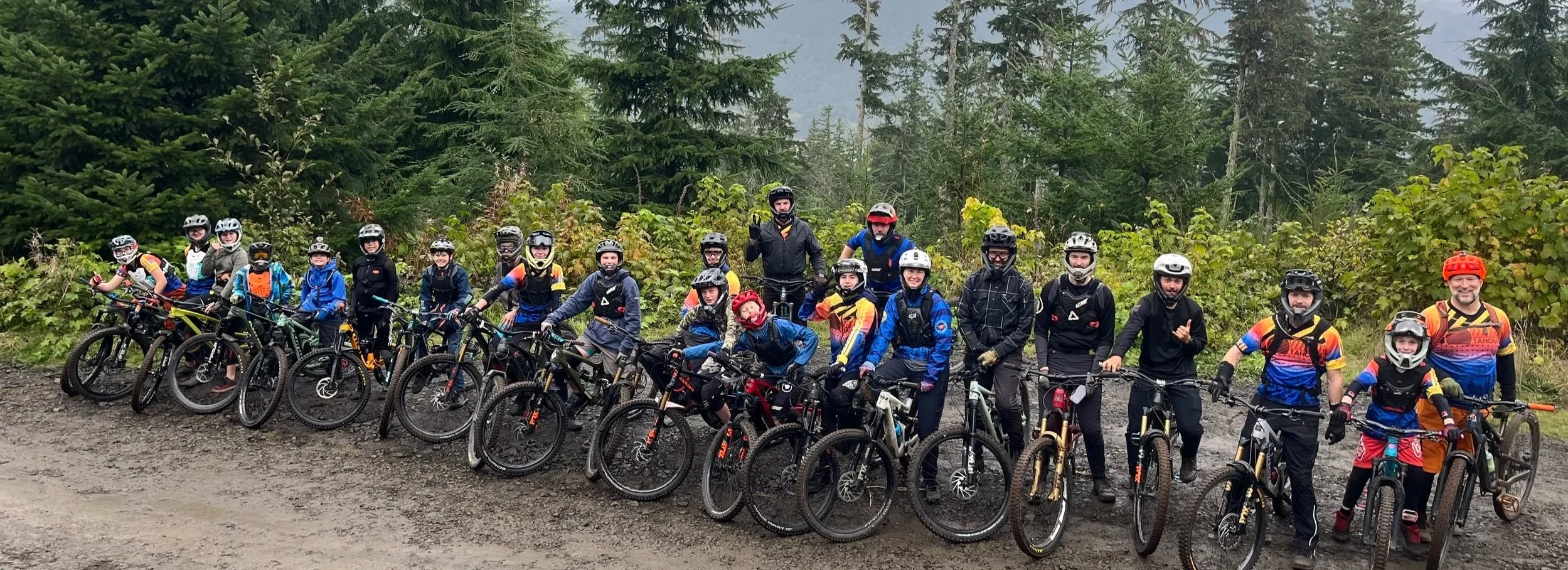 A large group of children and adults in biking gear and helmets, posing with their mountain bikes on a dirt trail in a forested area with green trees and cloudy sky.