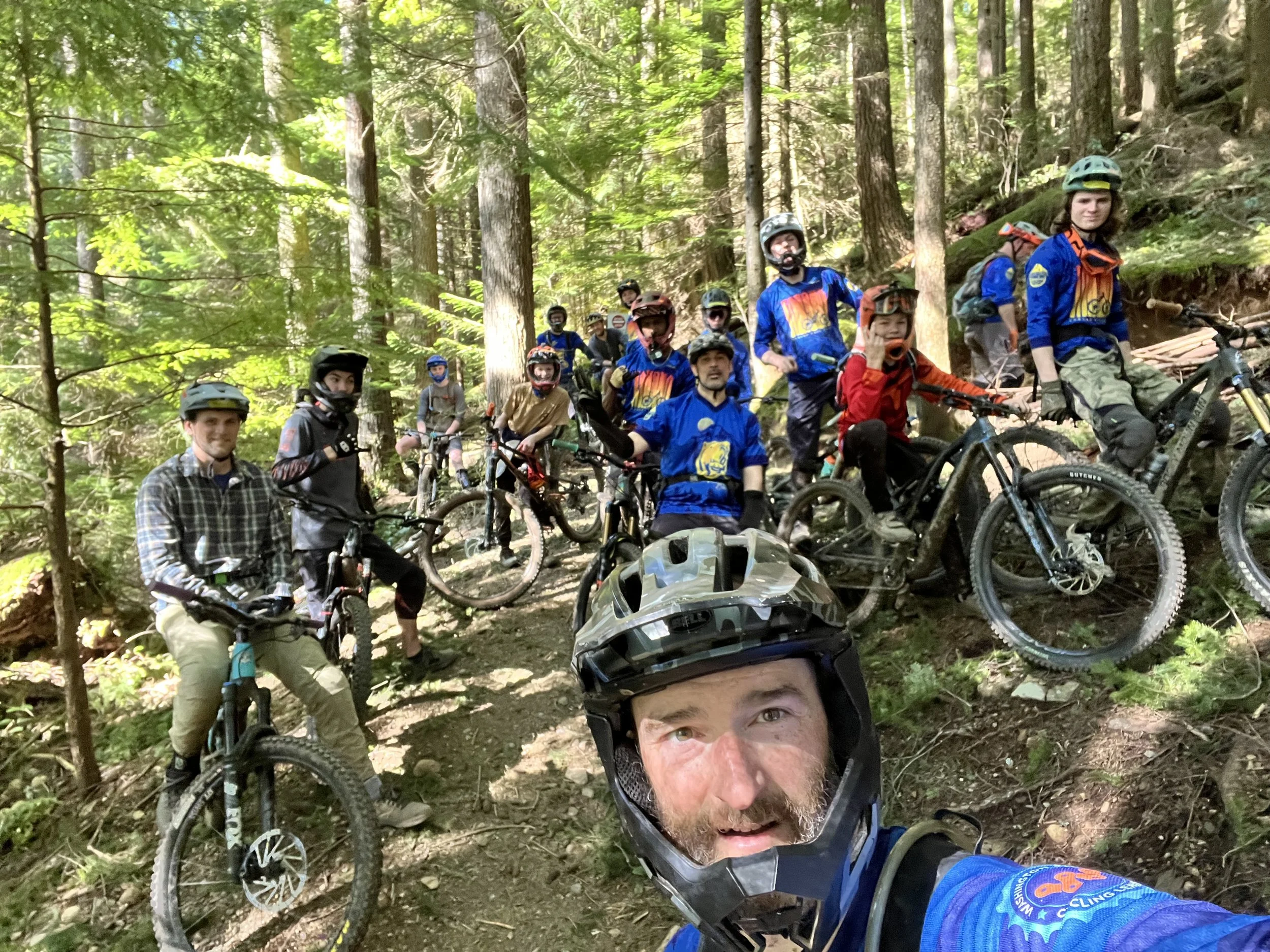 A group of mountain bikers, including children and adults, wearing helmets and outdoor gear, take a break on a forest trail with tall trees and green foliage.