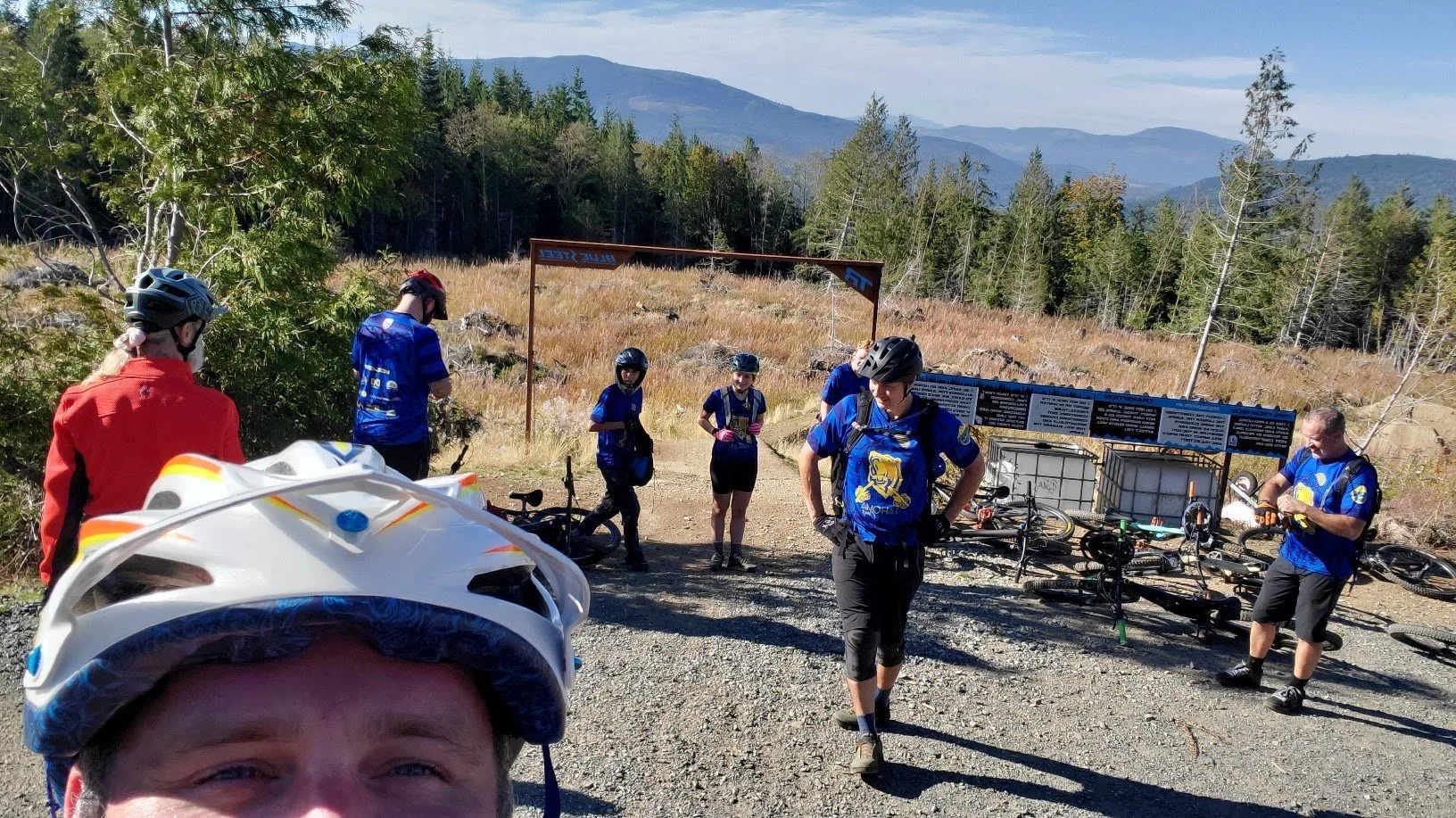 A group of mountain bikers in blue jerseys and helmets gathered on a gravel trail in a mountain forest. Some are talking while others are preparing their bikes, with a person taking a selfie in the foreground. The background features trees, mountains, and a blue sky.