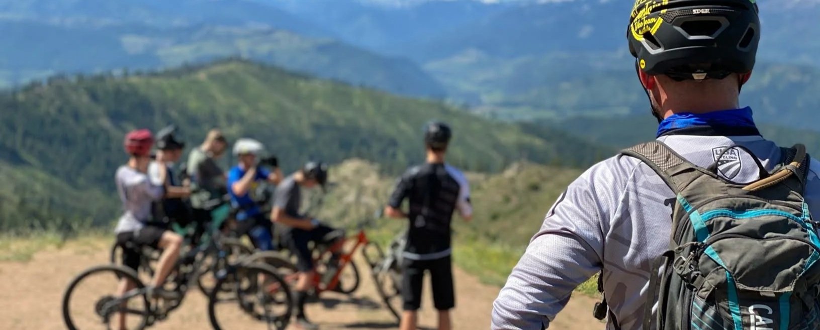 A group of mountain bikers on a trail with a scenic mountain landscape in the background; one cyclist in the foreground wearing a helmet and backpack, observing the others.