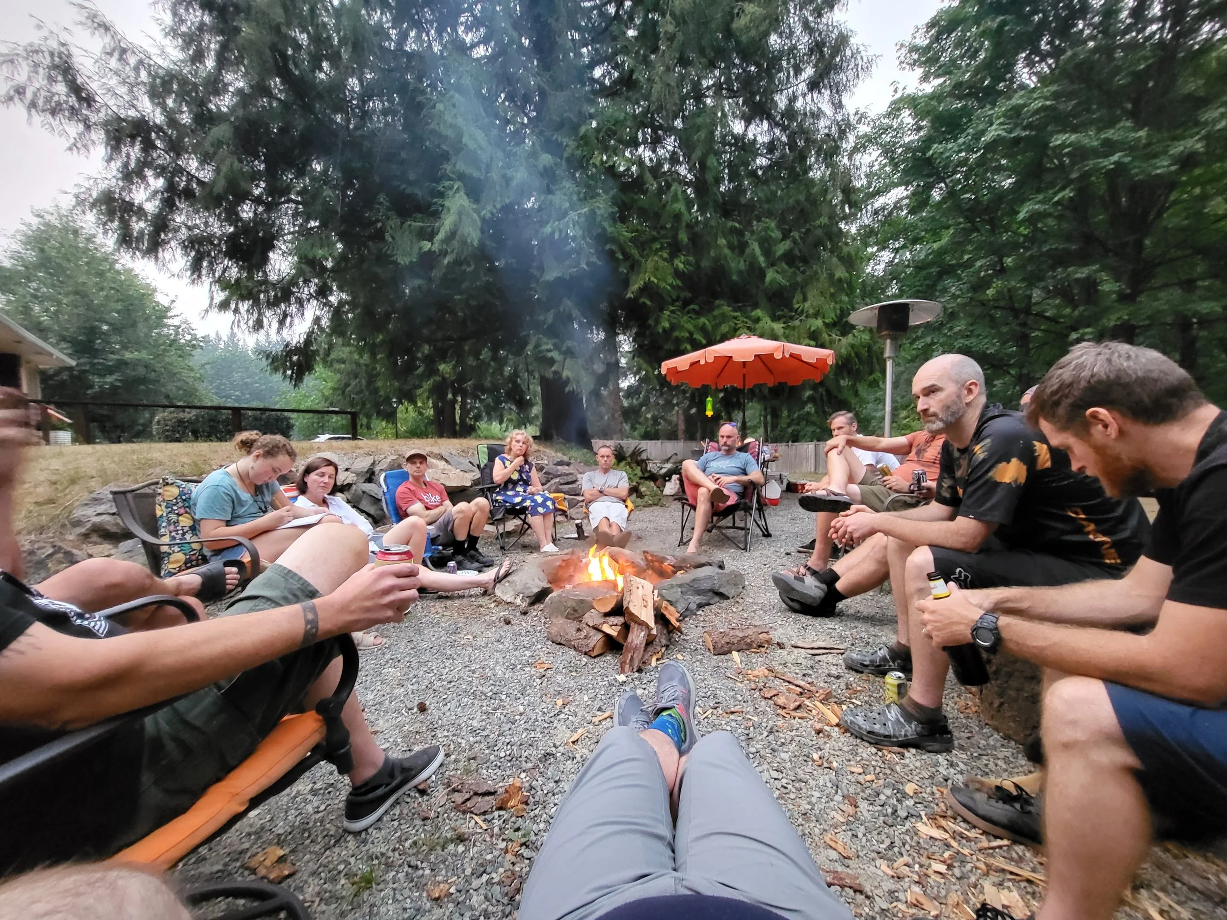 People sitting around a campfire outdoors in a wooded area, some holding drinks, with chairs and umbrellas visible.